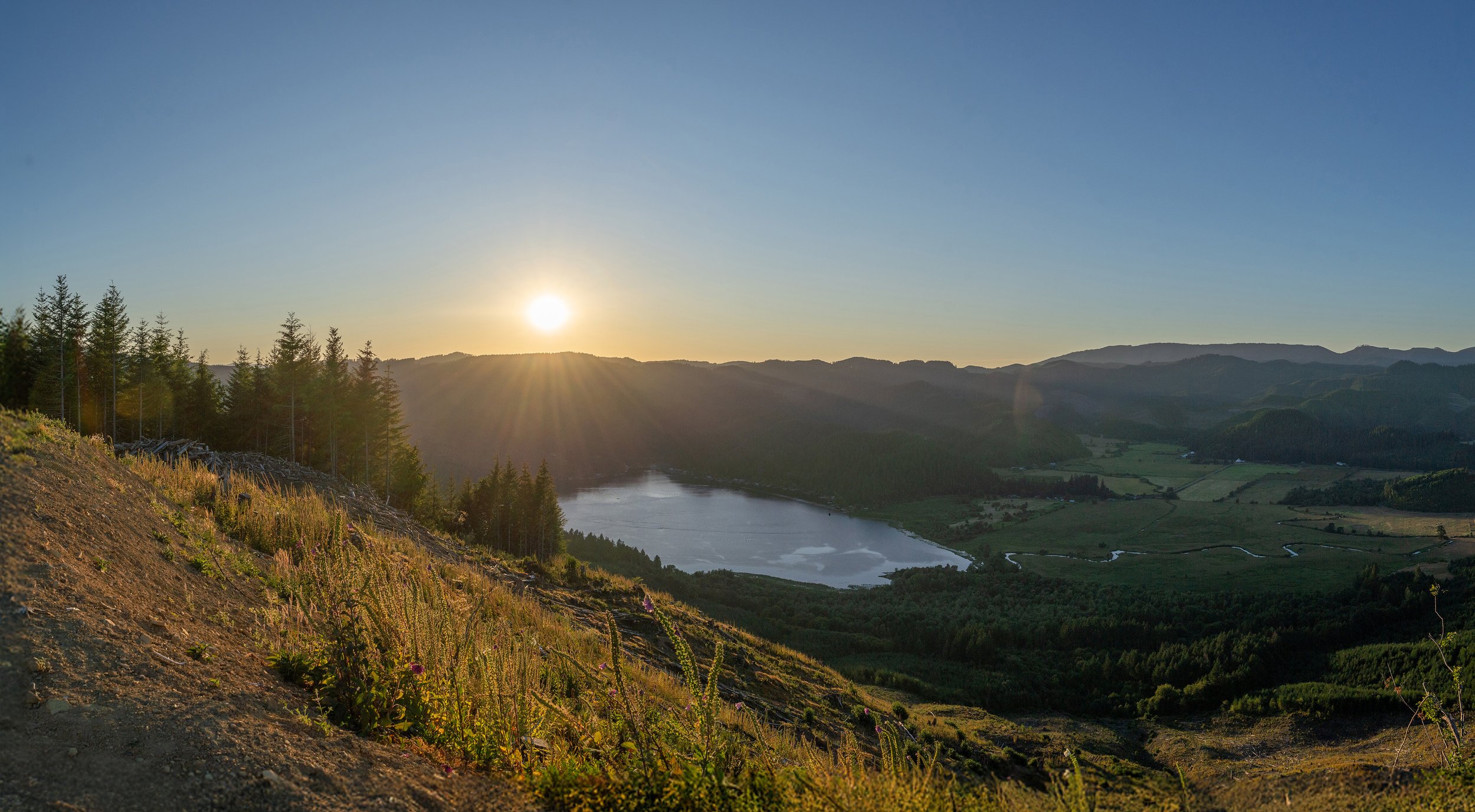 A scenic landscape at sunset showing a mountain range, a lake, and green fields with trees in a valley.