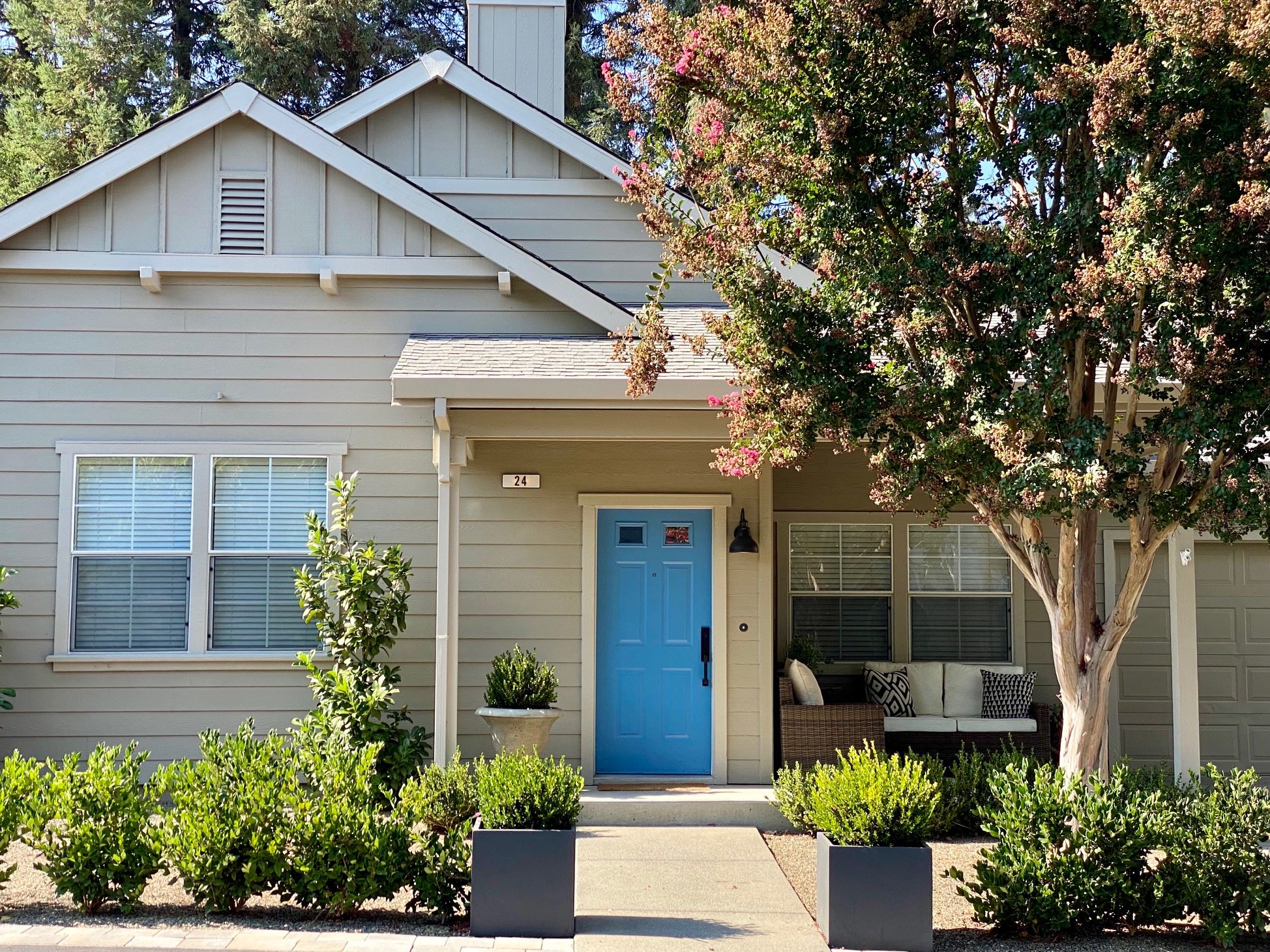 Front view of a house with beige siding, a front porch with a wicker sofa and cushions, a blue front door, potted plants, and a large tree with pink flowers in the yard.