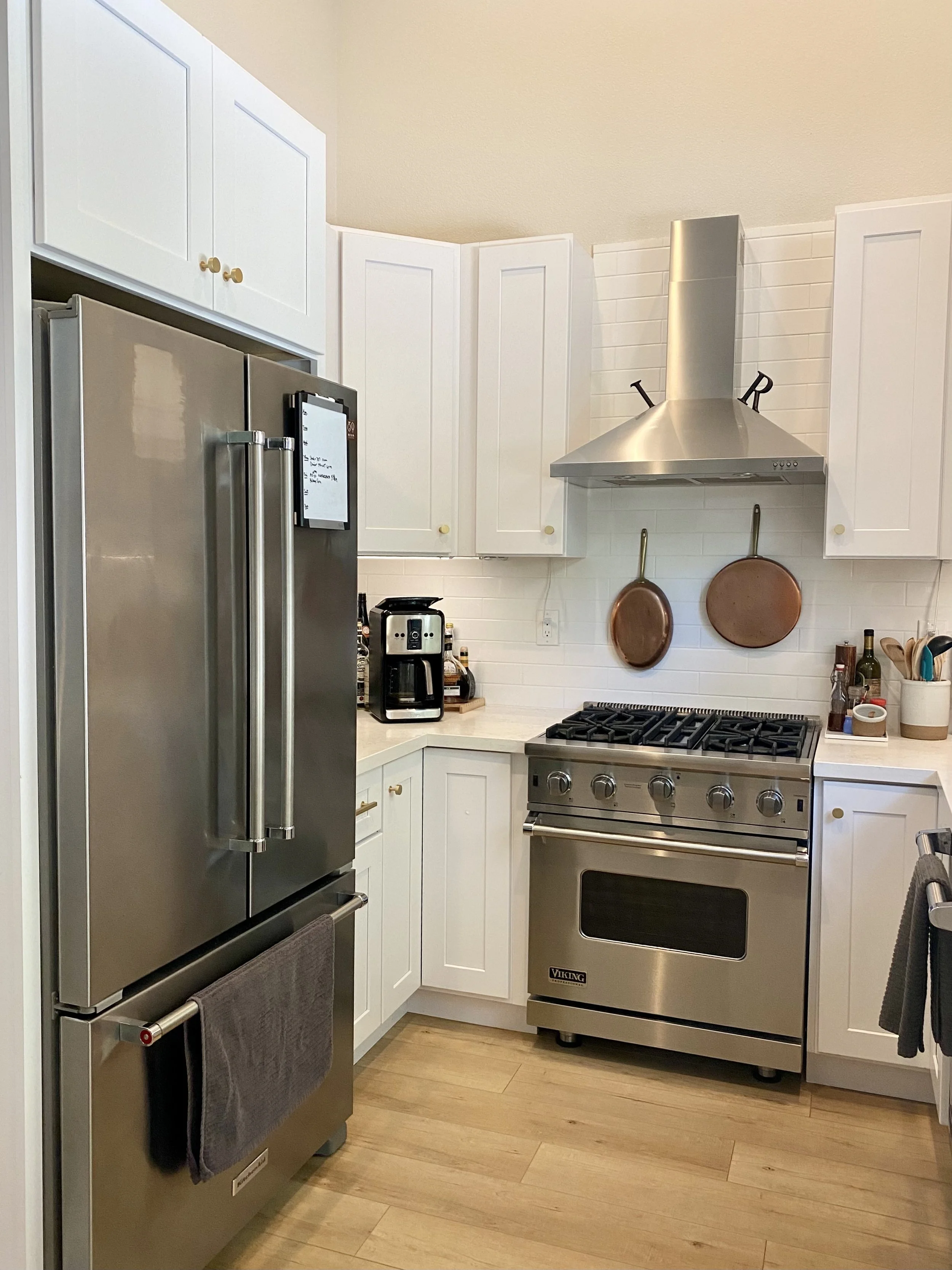 Modern kitchen with stainless steel refrigerator, oven, and range hood, white cabinets, copper pans, and a coffee maker.