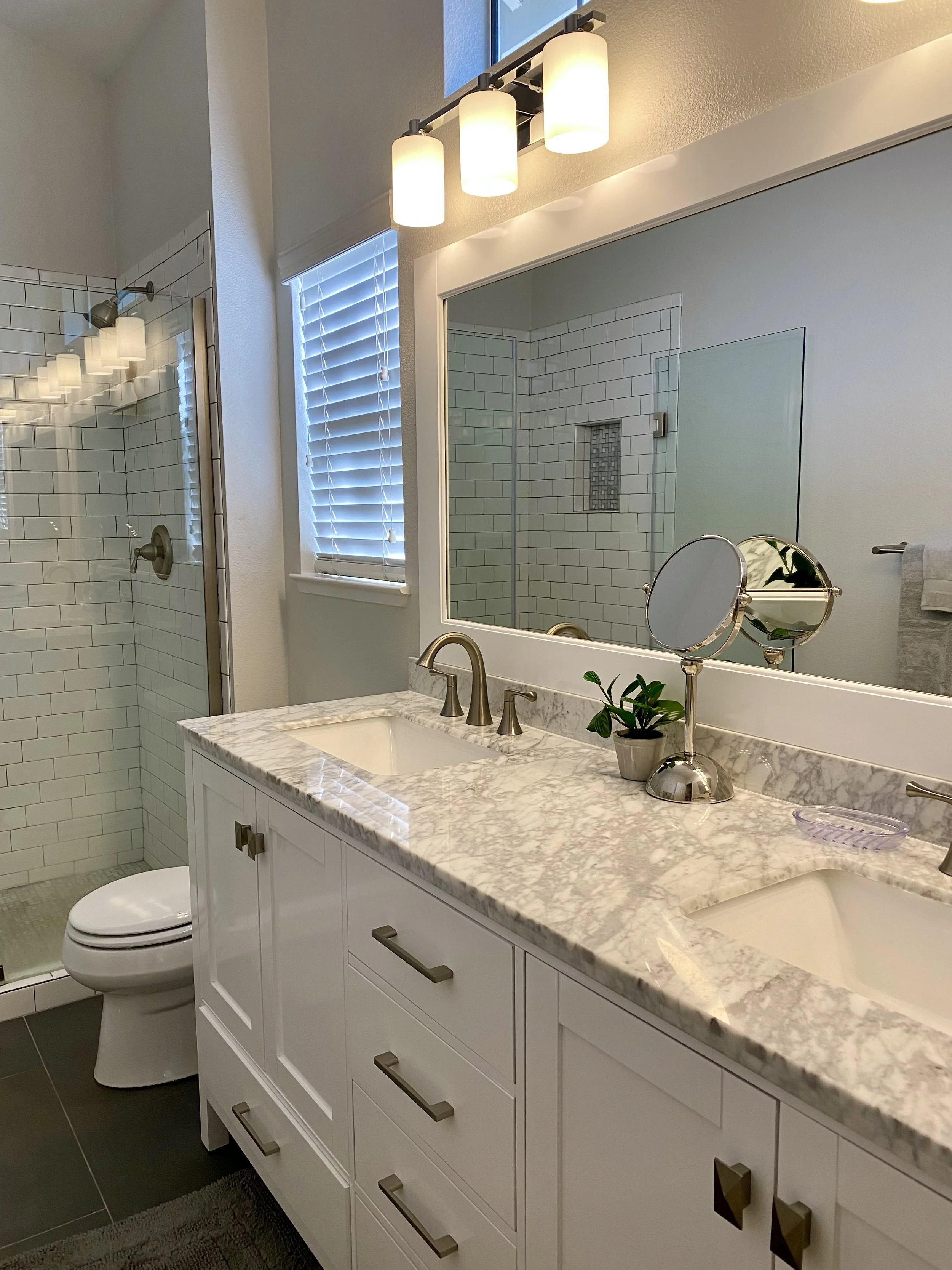 Bathroom with a marble countertop double sink vanity, a large mirror, a potted plant, a makeup mirror, and a window with blinds. A walk-in shower with glass panels and white subway tiles is visible in the background.