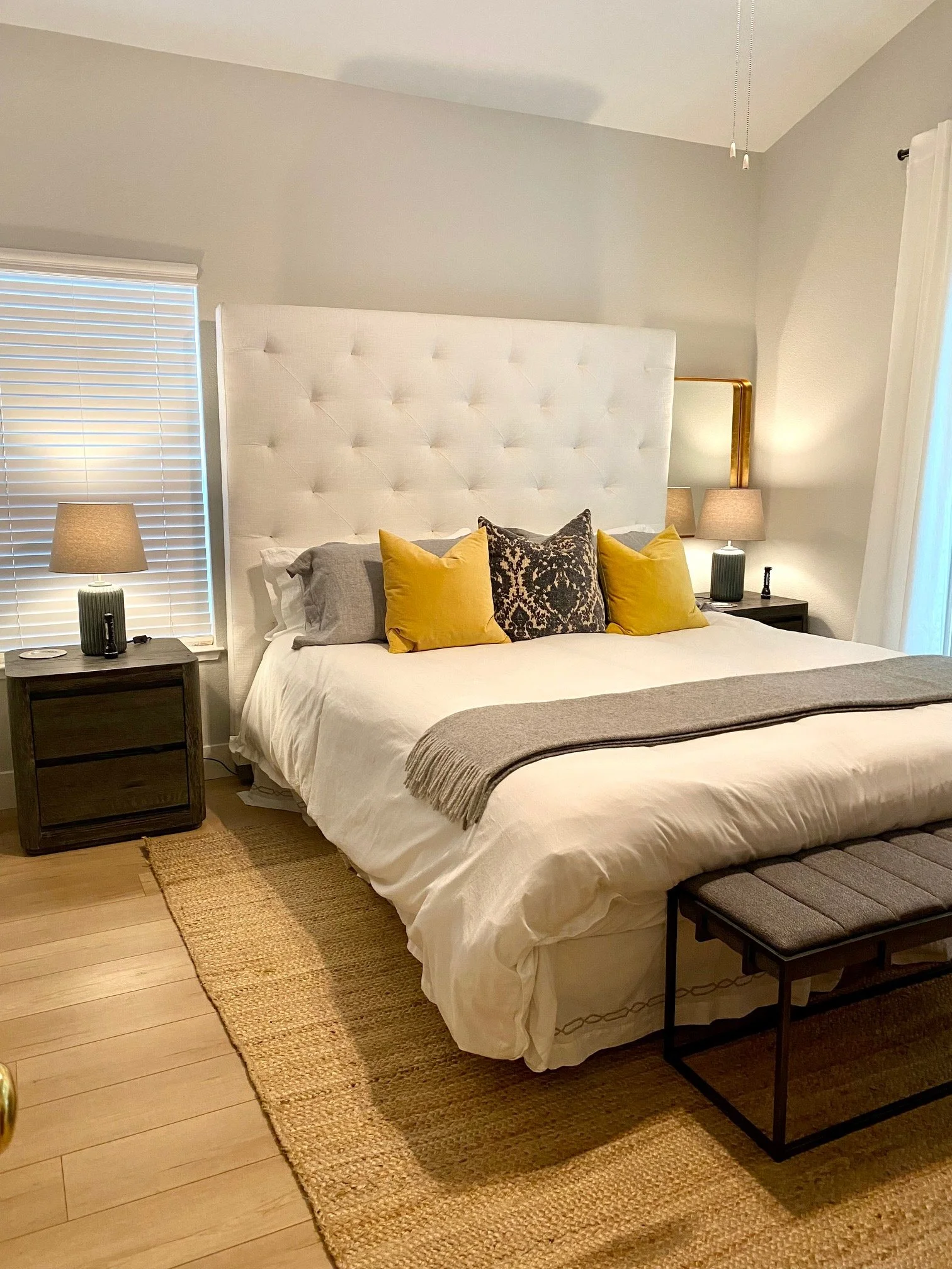 A bedroom with a large white tufted headboard, two matching nightstands with lamps, a bench at the foot of the bed, and a rug on wooden floor. There are decorative pillows on the bed.