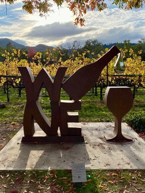 Sculpture spelling 'WINE' with creative designs; the letter 'I' is a wine bottle pouring into a glass, all set outdoors in a vineyard with mountains and a cloudy sky in the background part of the Art Walk in Yountville, California. 
