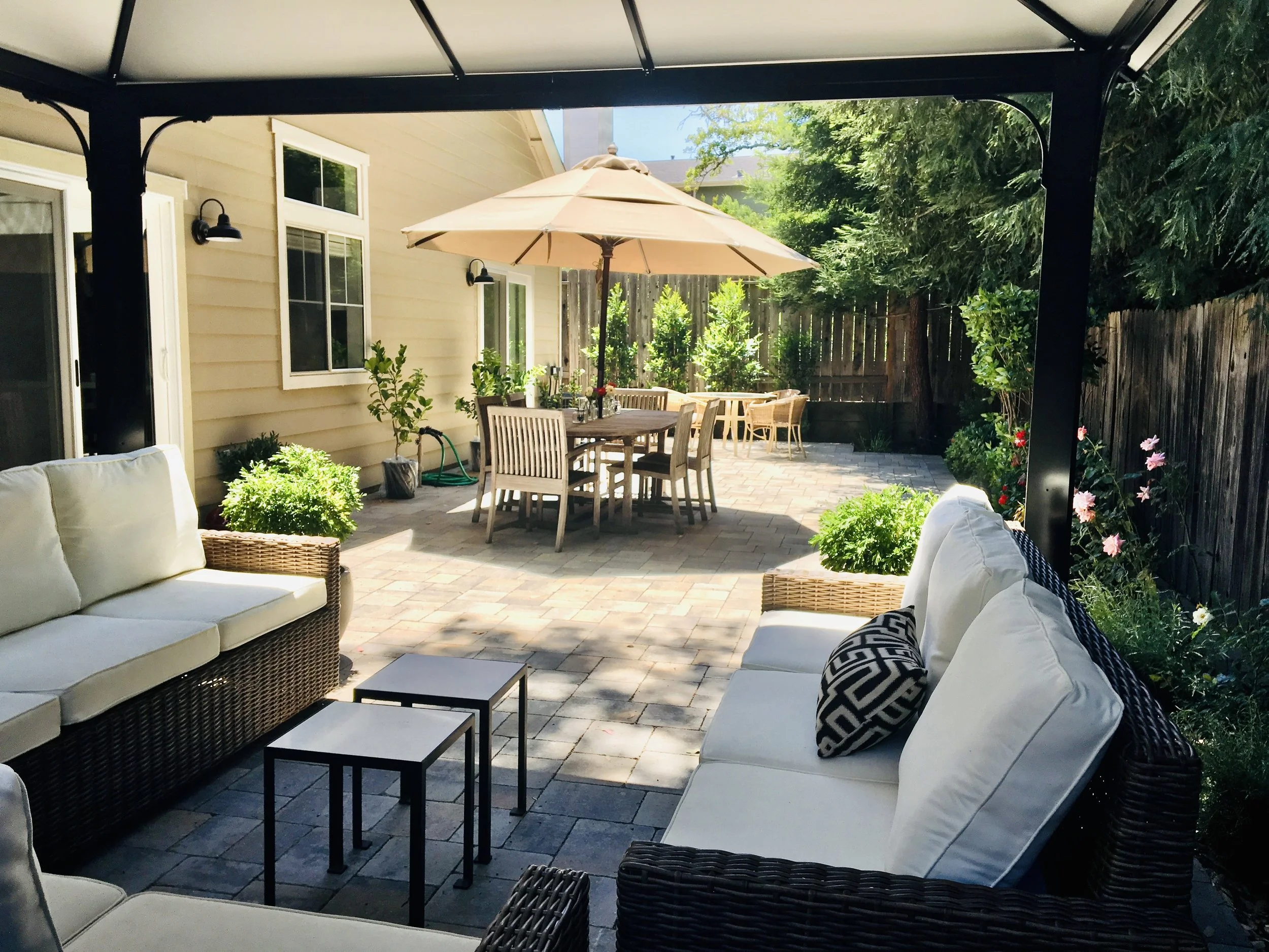 Outdoor patio with cushioned wicker furniture, potted plants, a dining area with a large umbrella, and a wooden fence surrounded by trees and flowers.