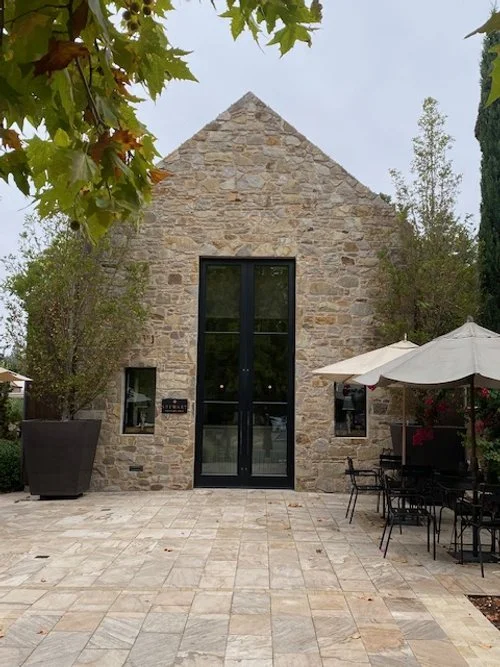 Stewart Cellars winery. Stone building with large glass doors, outdoor seating area with tables and chairs, umbrellas, trees, and a cloudy sky.