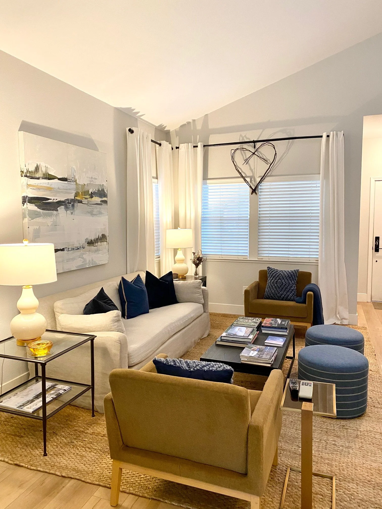 Living room with beige sofa, green armchair, blue ottomans, black coffee table with magazines, side table with lamps, and abstract wall art, with heart-shaped wall decor above window