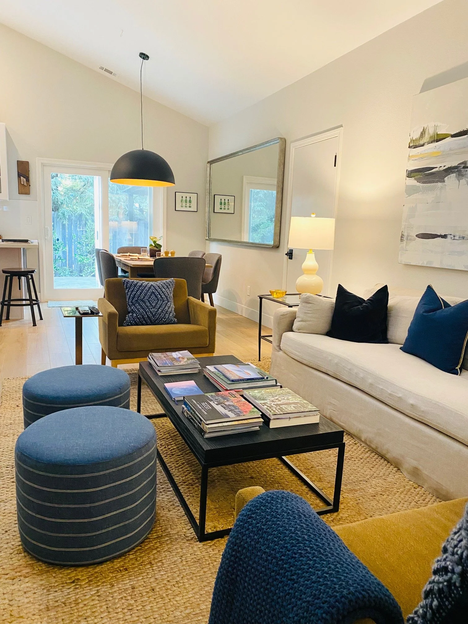 Living room with beige sofa, yellow armchair, blue ottomans, black coffee table with magazines, large mirror, wall art, and a dining area with round table and four chairs, natural light from sliding door.
