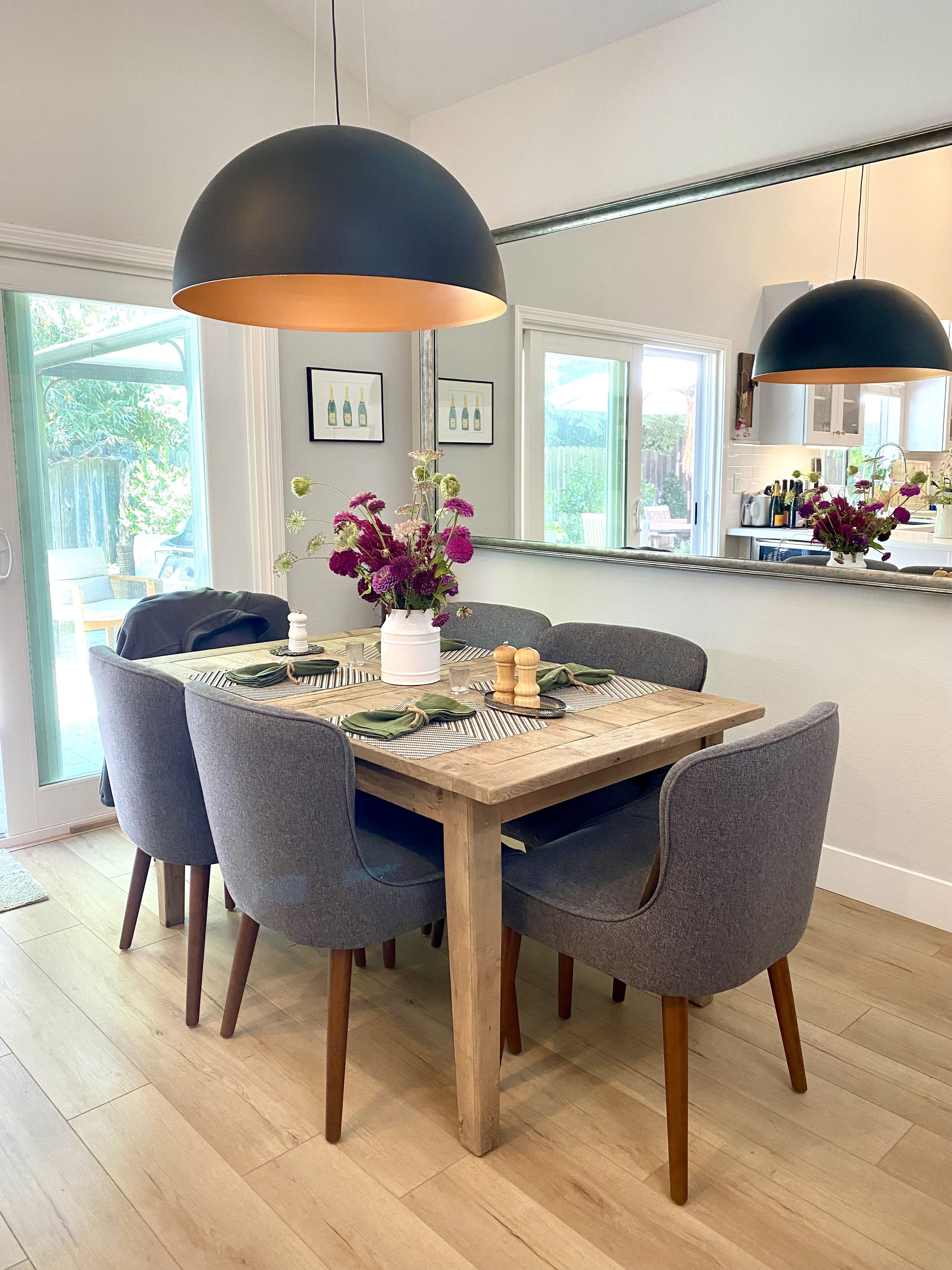 A dining room with a wooden table surrounded by six gray upholstered chairs with wooden legs. The table has a white vase with pink, purple, and white flowers and place settings with green napkins. Black pendant lights hang above the table. There is a large mirror on the wall reflecting part of the room and a sliding glass door leading outside.