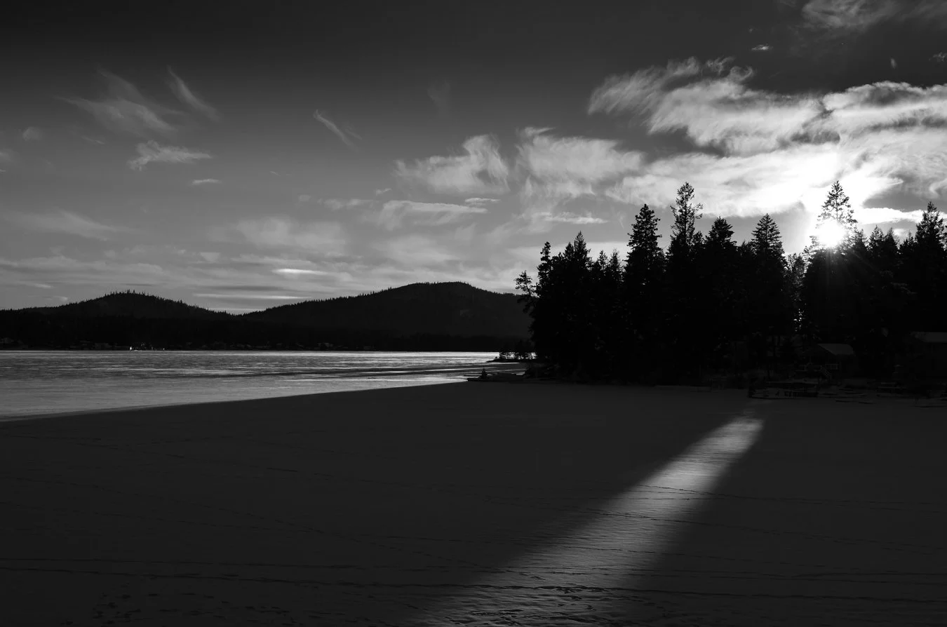Black-and-white photo of a lakeside with a shadowed dock, a group of trees near the water, distant hills, and a sky with wispy clouds, with the sun low on the horizon.