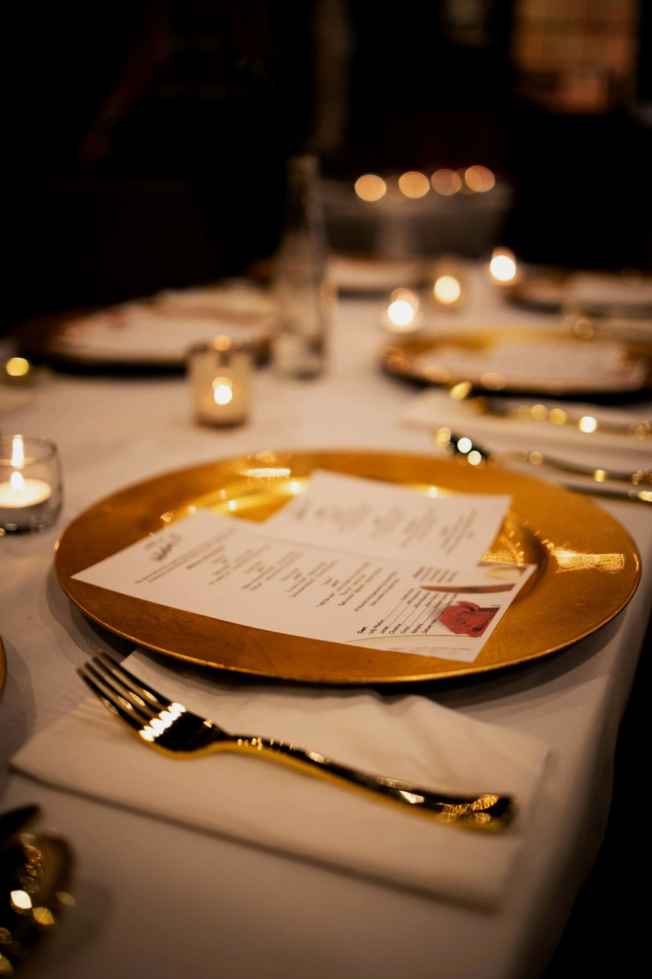 Ein eleganter Tisch mit goldener Platte, einem Menü, einem Messer und einer Gabel, umgeben von Kerzenlicht bei einer festlichen Veranstaltung.