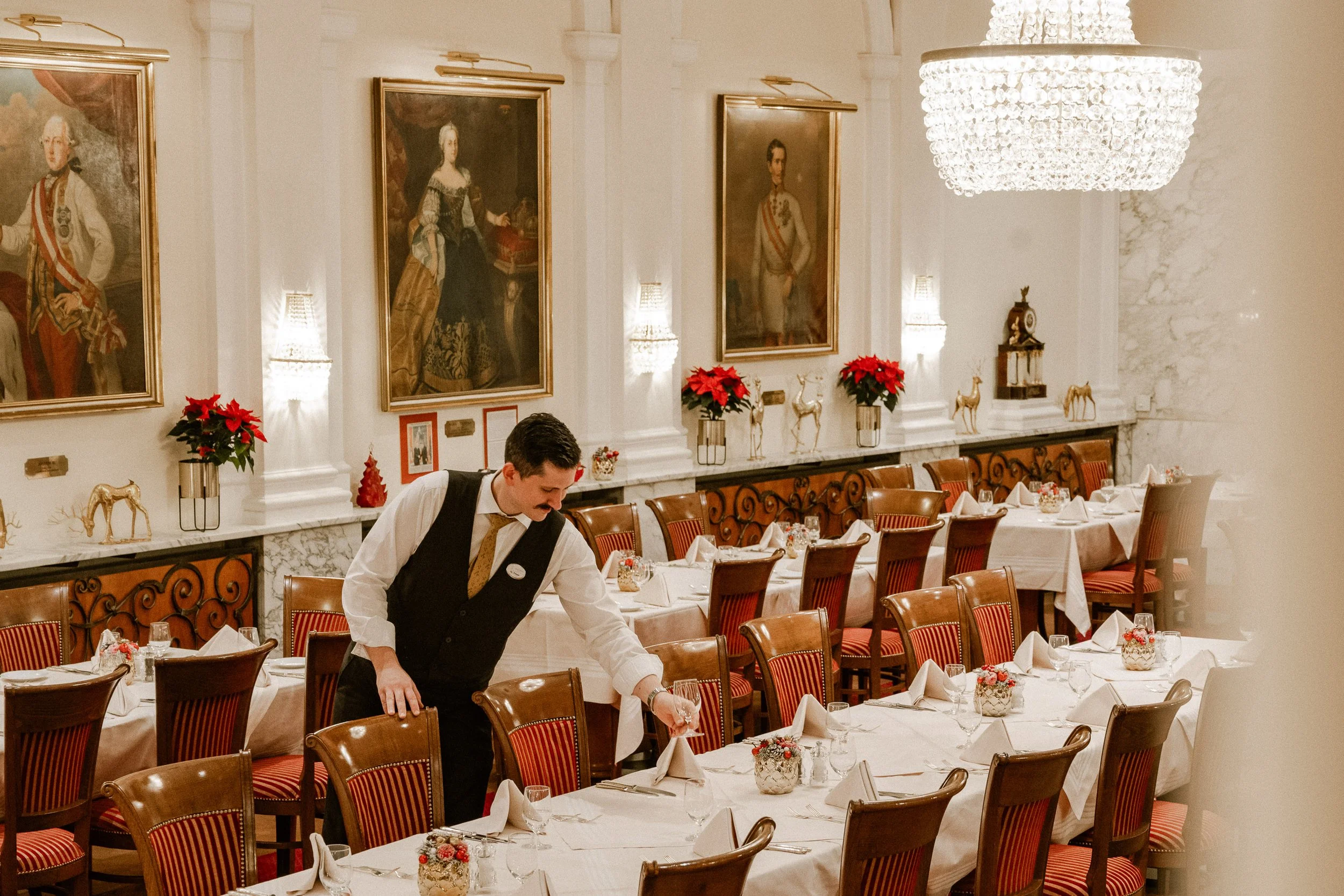 Ein festlich gedeckter Speisesaal mit weißen Tischdecken, rot karierte Stühlen, Weihnachtsdekorationen und einem Kellner, der einen Tisch deckt, große Gemälde an der Wand und ein Kronleuchter an der Decke.