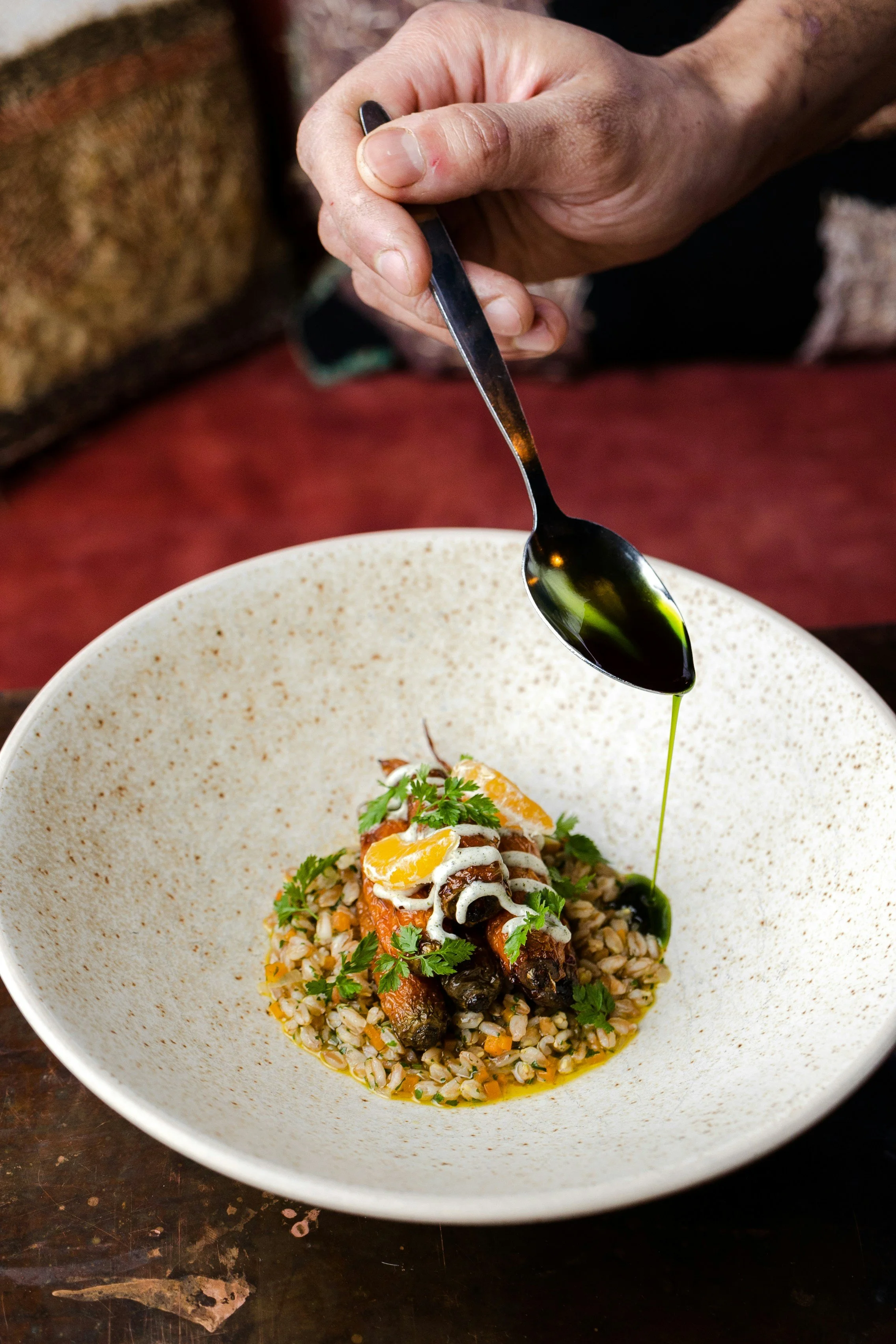 Ein Teller mit Gericht aus Reis, Gemüse, Fleisch und einer Rolle, garniert mit frischen Kräutern, während eine Person mit einem Löffel grüne Soße über das Gericht gießt.