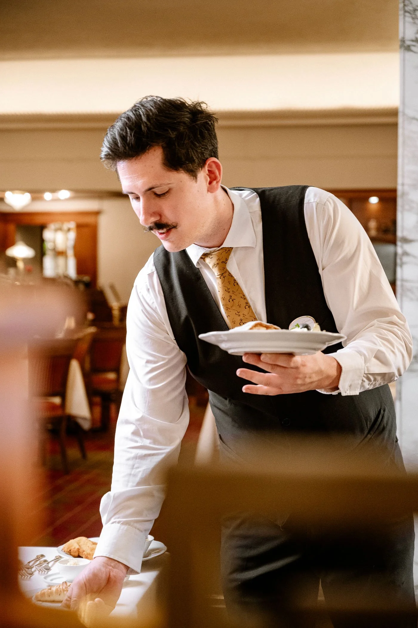 Ein männlicher Kellner in einem weißen Hemd, dunkler Weste und gelbem Schlips, serviert Speisen in einem Restaurant.