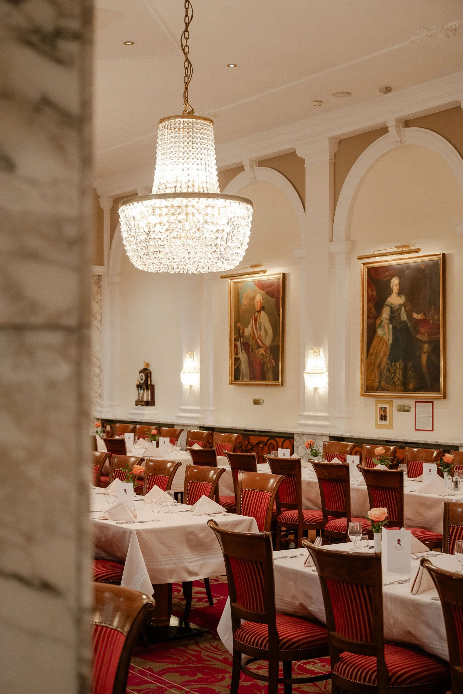 Ein eleganter Speisesaal mit langen Tischen, weißen Tischdecken, Blumen, Kristallkronleuchtern und Gemälden an den Wänden.