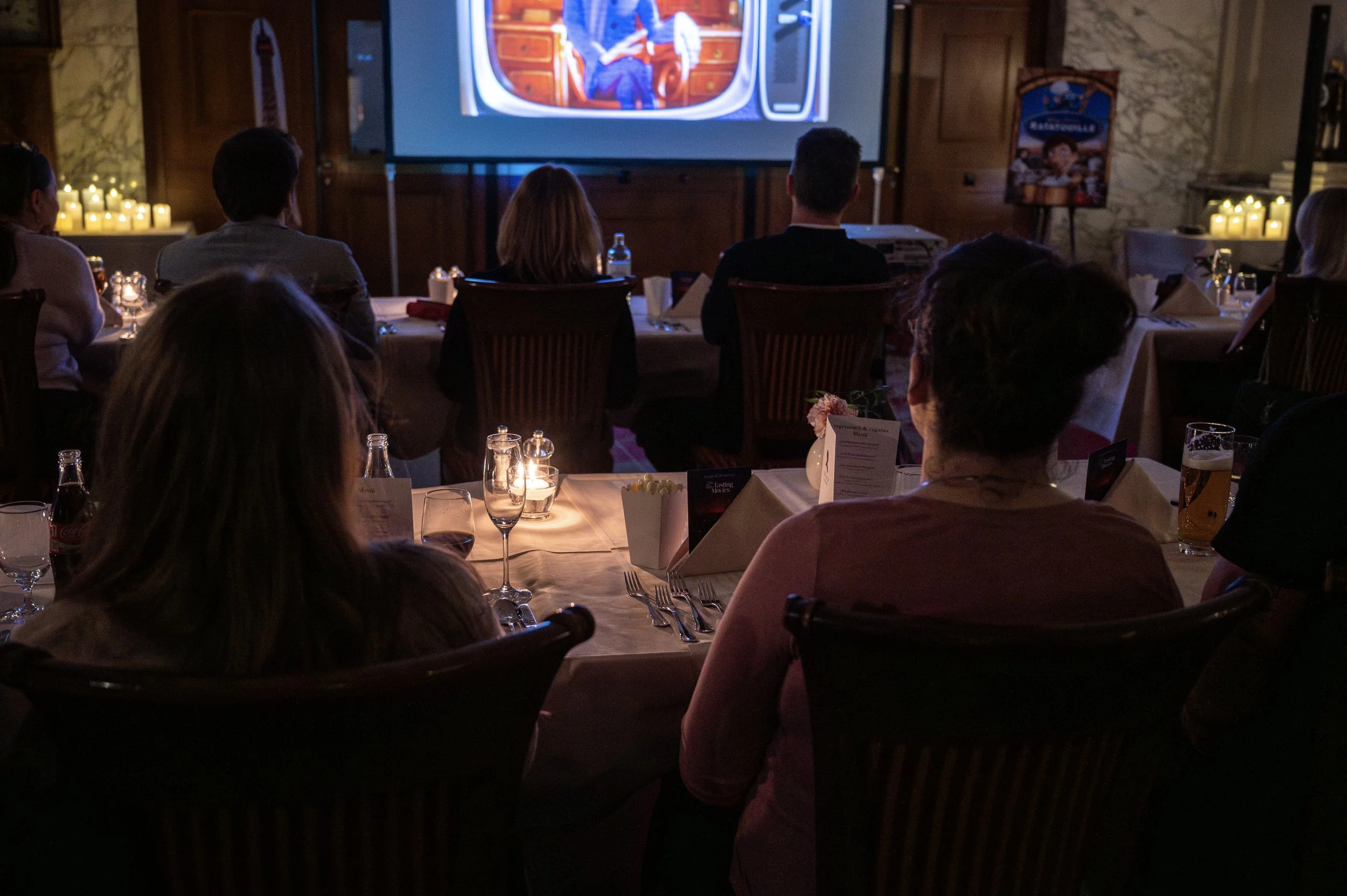 Menschen sitzen in einem dunklen Raum und schauen einen Fernseher, auf dem eine Sendung läuft.