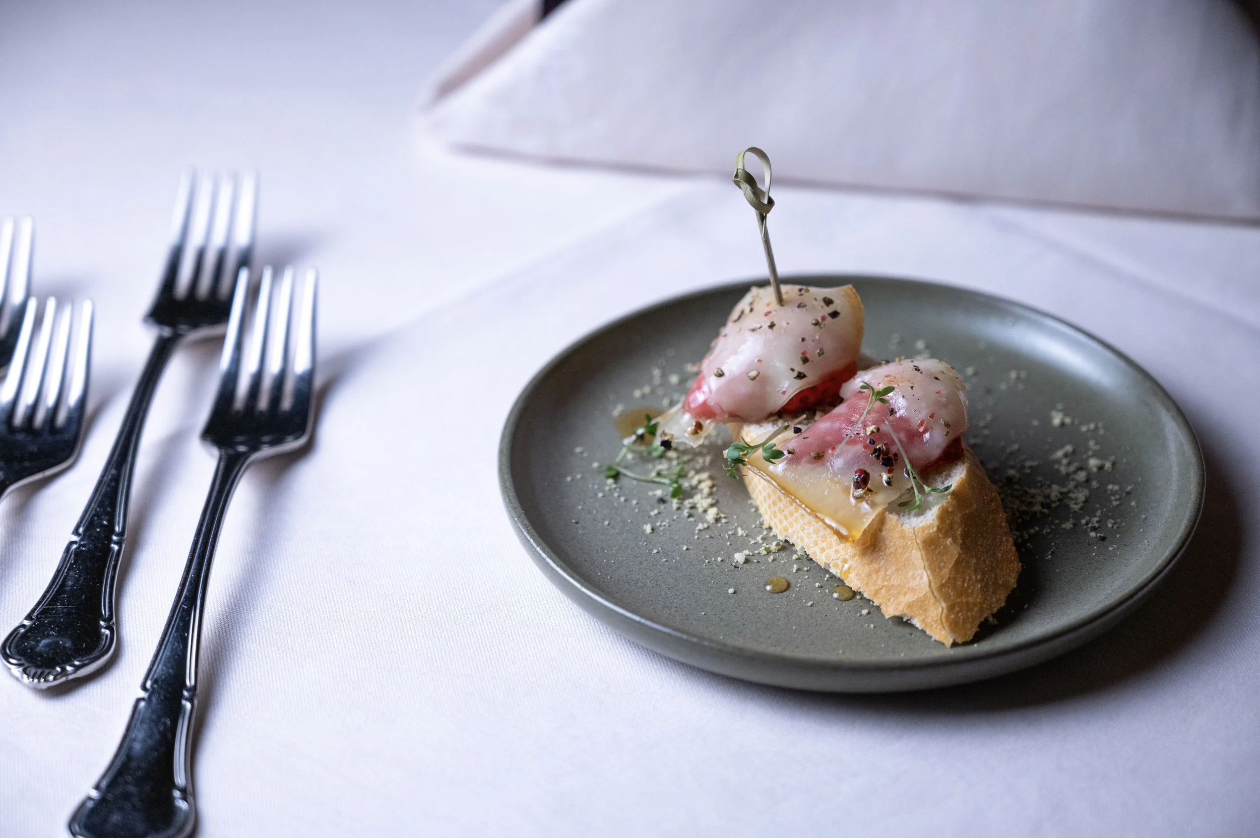 Eine kleine Portion Garnelen auf geröstetem Baguette, garniert mit Kräutern, Pfeffer und Öl, serviert auf einem grauen Teller.