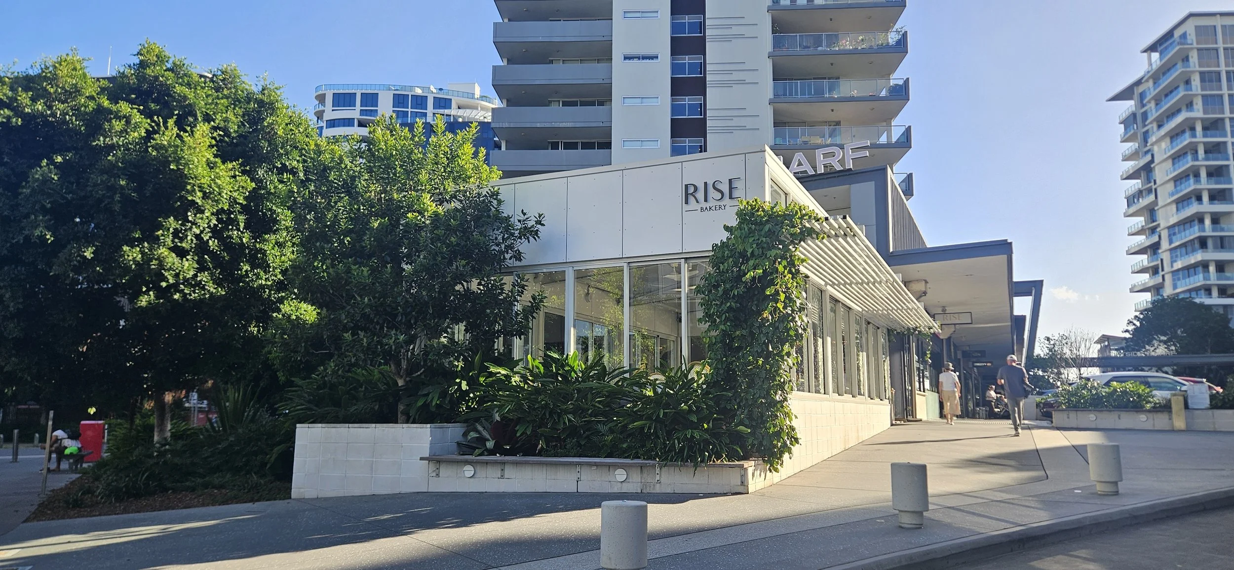 Street view of a modern building with a bakery called 'Rise Bakery' on the ground level, surrounded by trees and greenery, with high-rise residential buildings in the background on a sunny day.