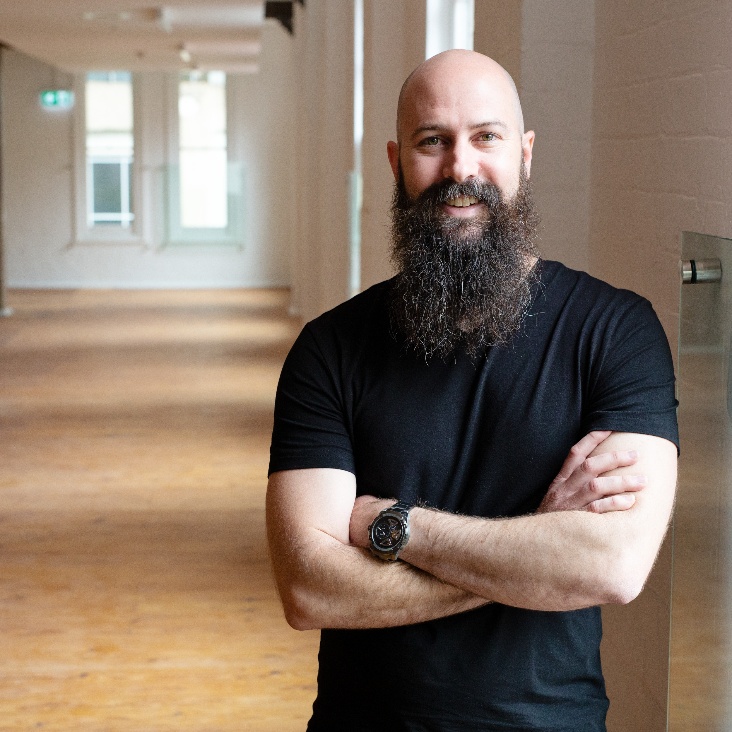 A man with a shaved head and a full beard smiling with arms crossed, standing indoors near a brick wall with windows in the background.