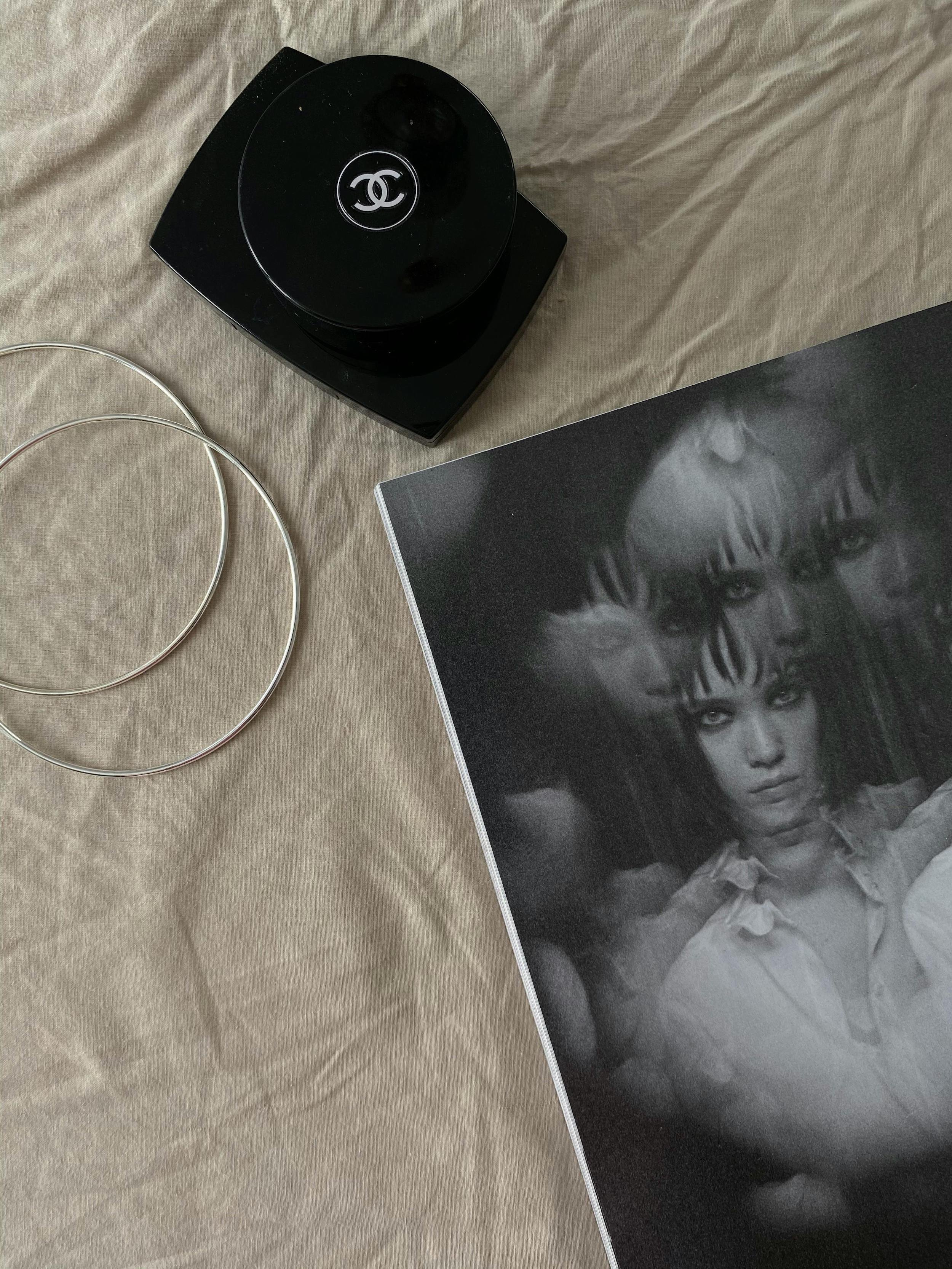 A black Chanel compact, two silver bangles, and a black-and-white photograph of three women with intense expressions, laid out on a beige fabric surface.