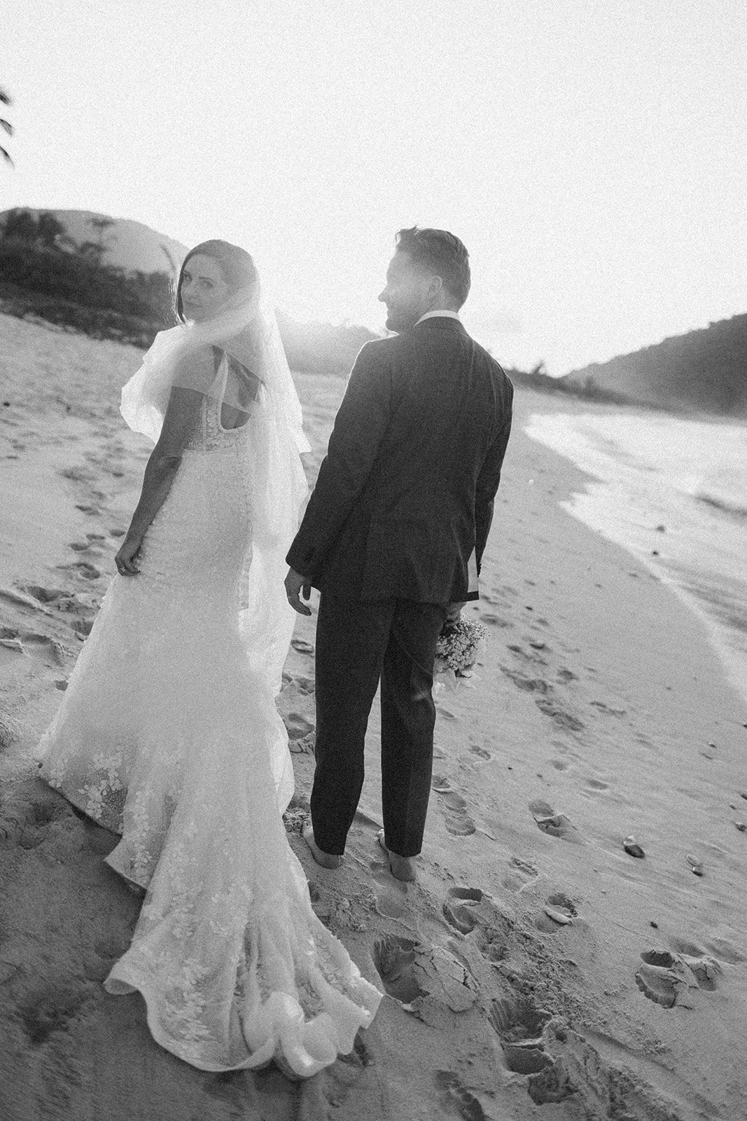 A black and white photo of a bride and groom on the beach at sunset, the bride wearing a wedding dress and veil, the groom in a suit, holding a bouquet, with footprints in the sand behind them.