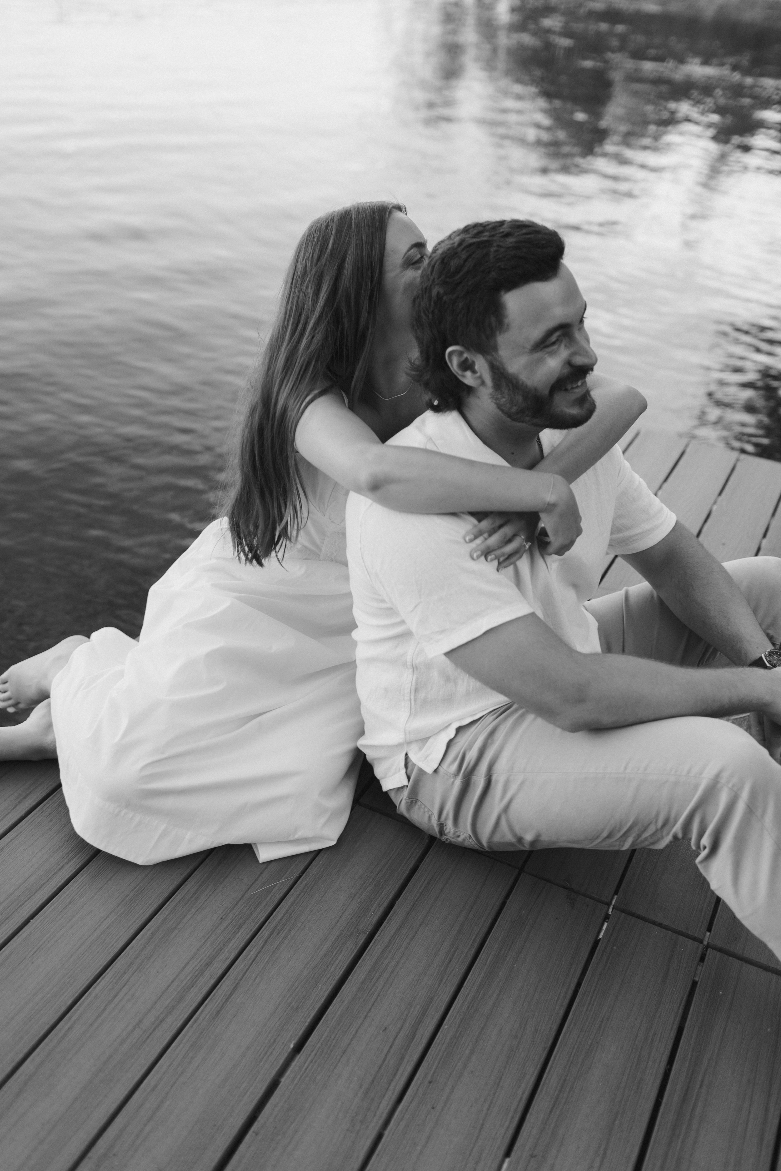 A black and white photo of a couple sitting on a wooden dock by the water. The woman is behind the man, hugging him and kissing his cheek. They are smiling and relaxed, enjoying each other's company.