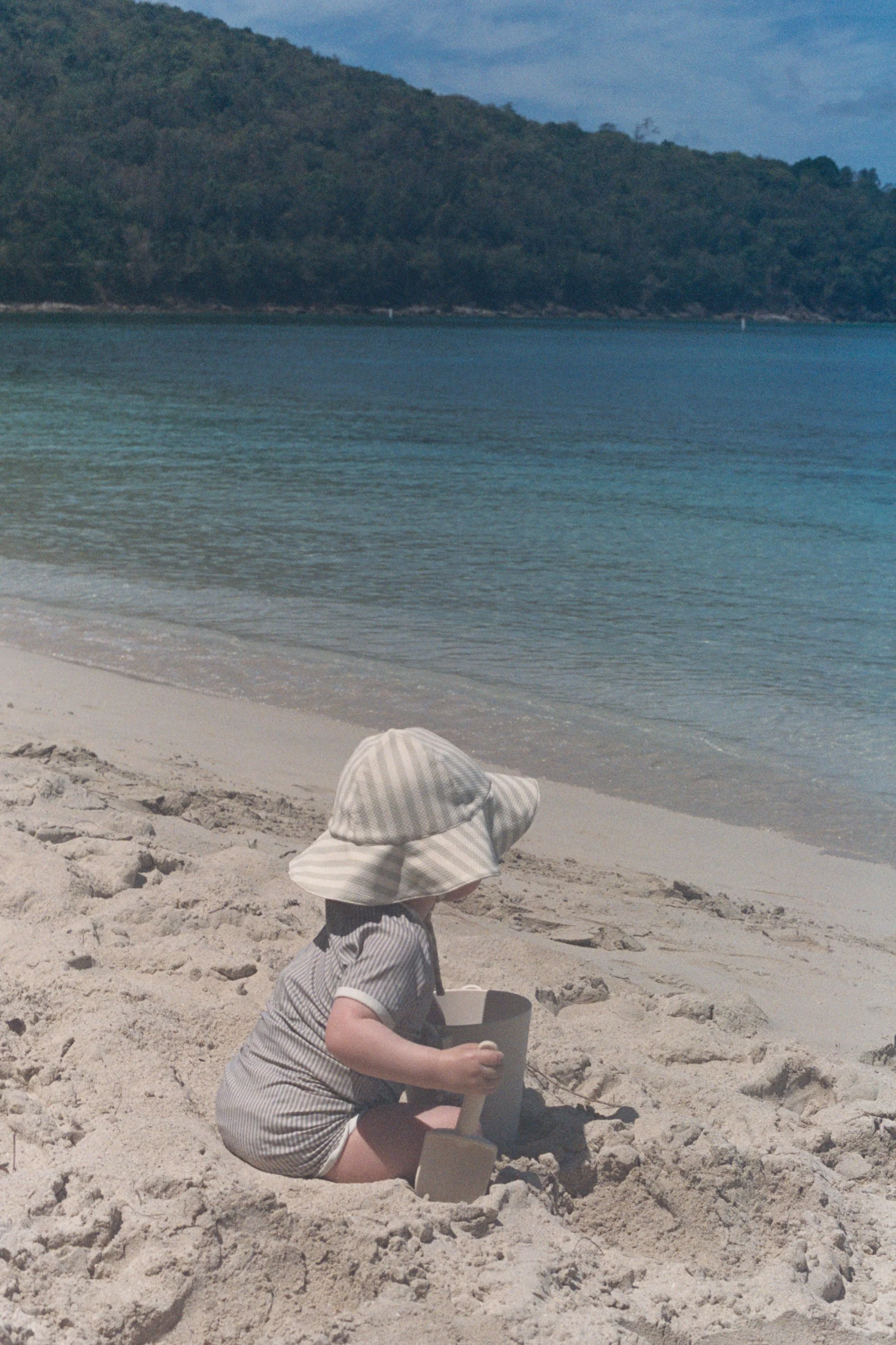 A young child sitting in the sand on a beach, wearing a striped sun hat and striped outfit, playing with a small bucket and shovel near the shoreline with calm water and a mountain in the background.