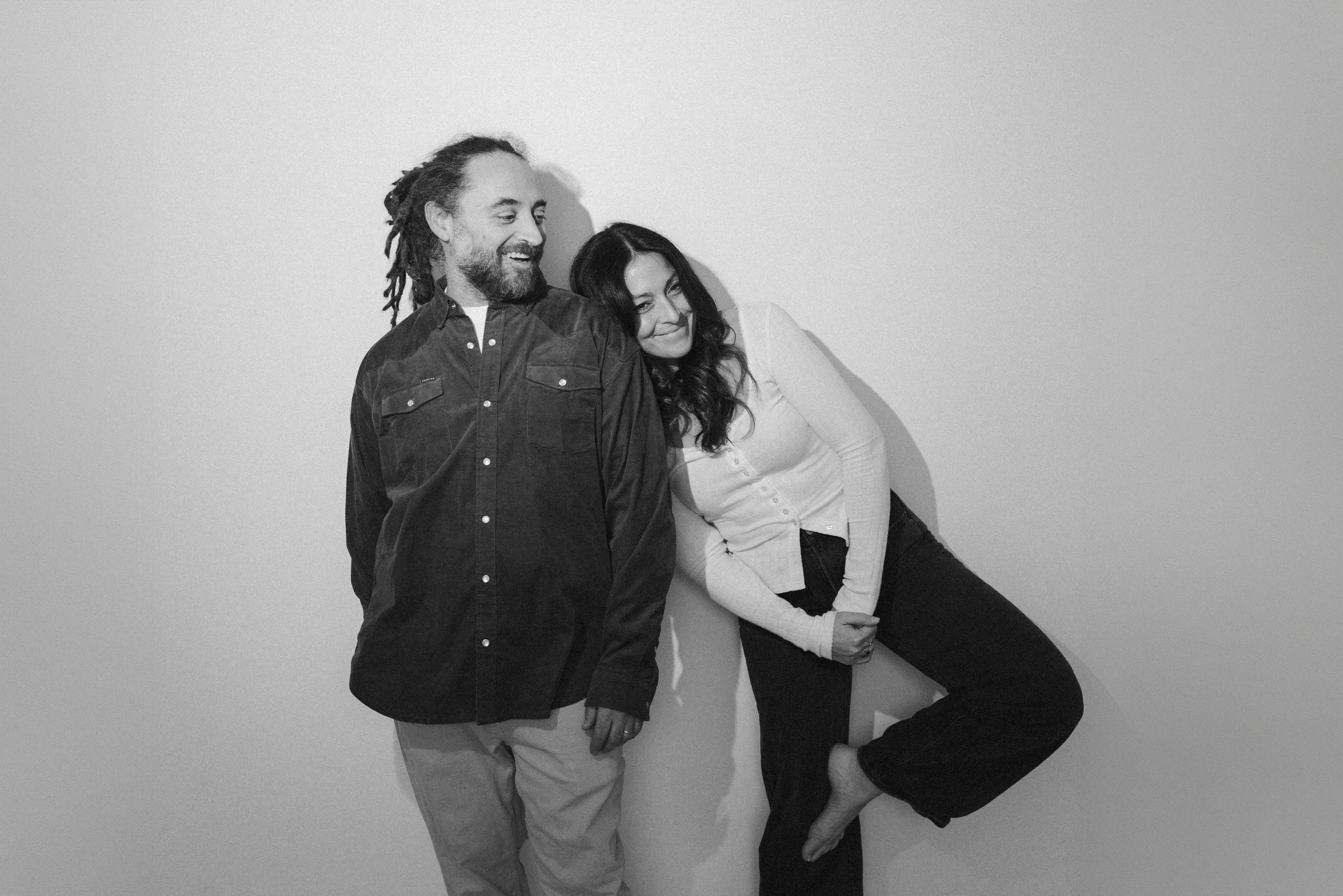 A man with dreadlocks and a beard, wearing a dark button-up shirt and light pants, standing next to a woman with long dark hair, wearing a light cardigan and dark pants, both smiling and leaning against a plain wall.