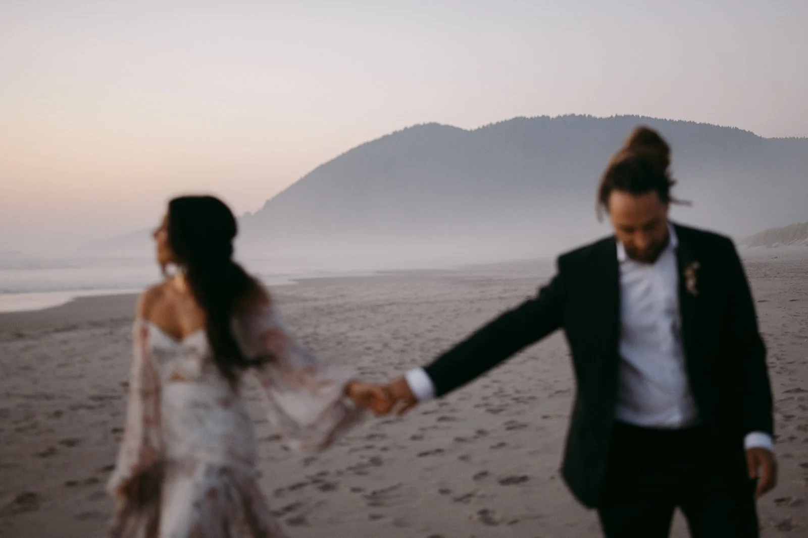 A man and woman holding hands on a beach at sunset, the woman in a flowing white dress and the man in a black suit, with a mountainous landscape in the background.
