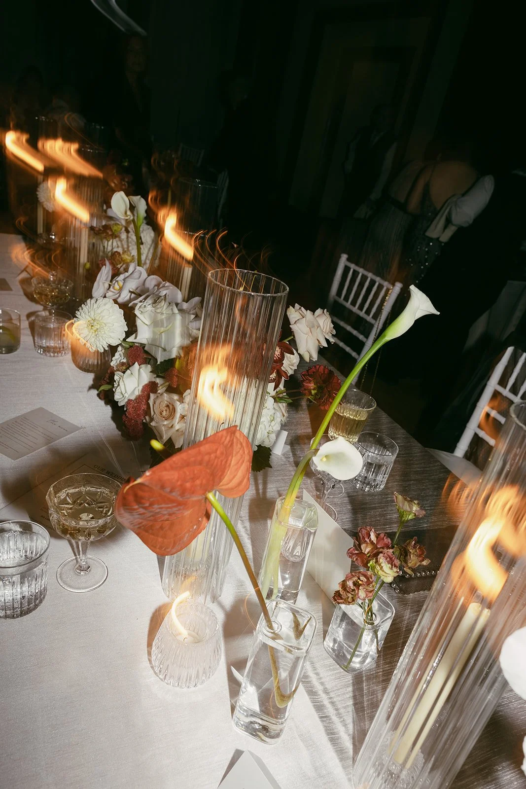 A decorated table with flowers and candles in vases, set for a formal event or celebration, with a blurred background of people in a dimly lit room.