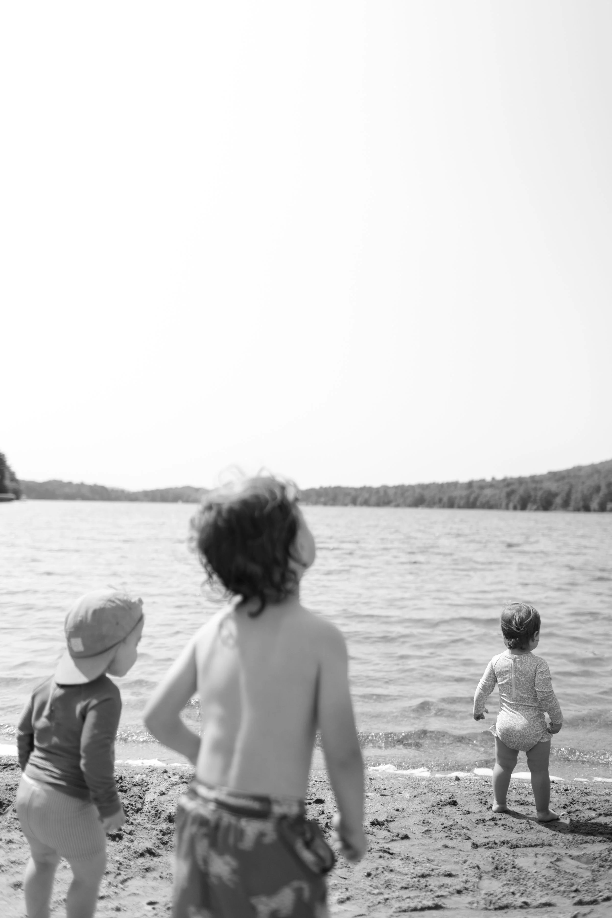 Three children on a sandy beach near water, with trees and hills in the background; one child is looking out at the water, two others are closer to the camera, one with back to the camera, the other partially visible, all in casual beach clothing, black-and-white photo.