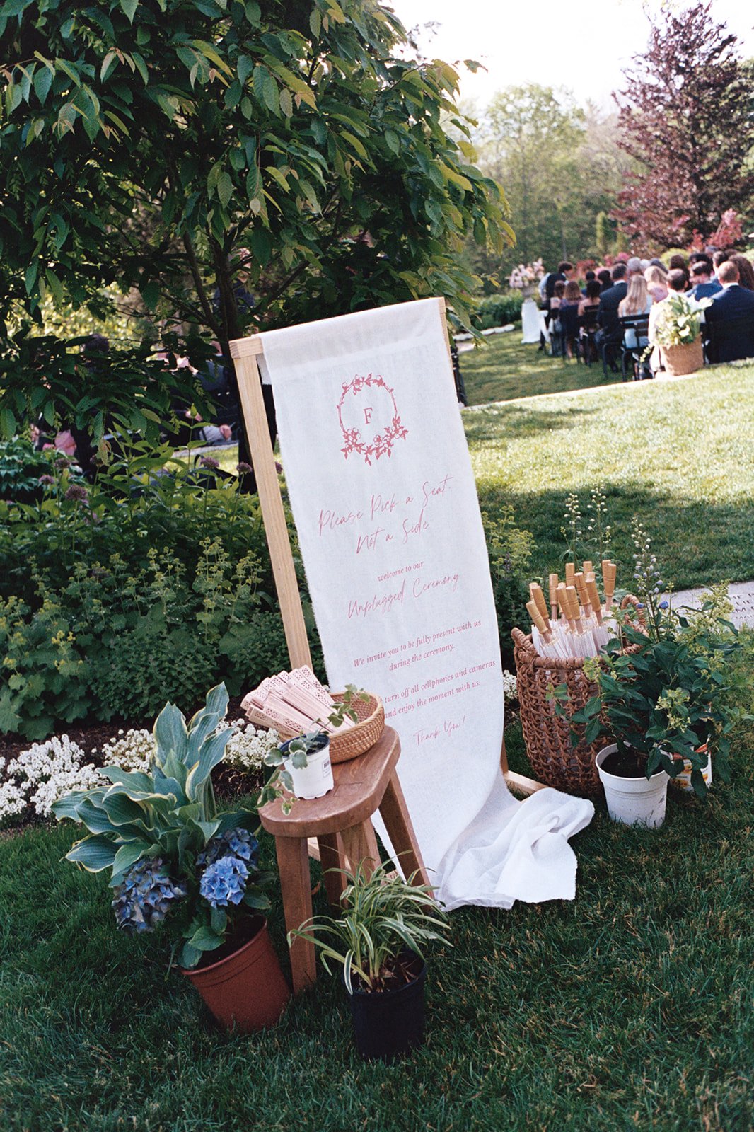 A wedding ceremony outdoor setup with a seated audience, a large announcement board with a floral monogram, plants, and a small table with pink paper fans.
