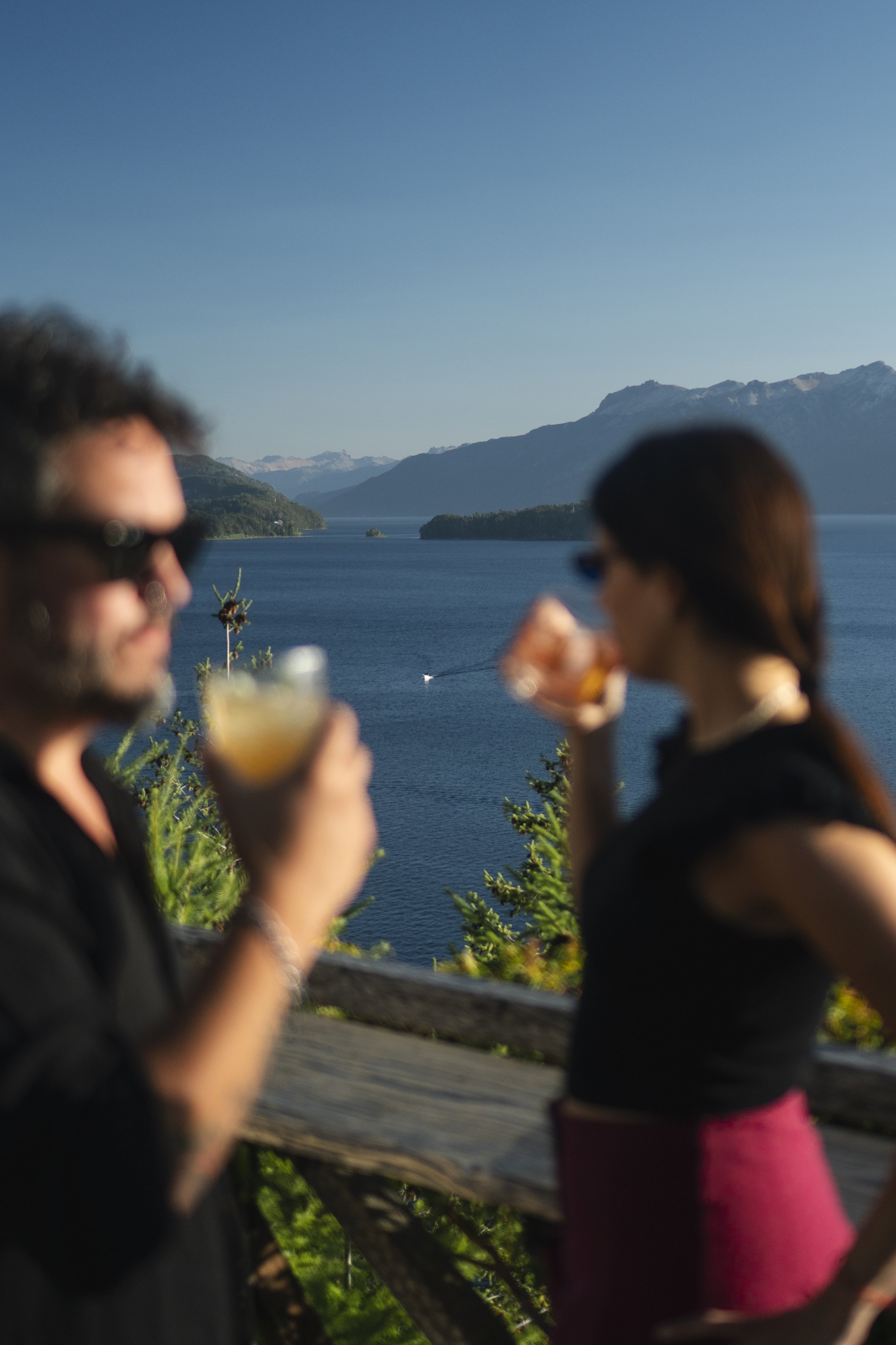 Dos personas con gafas tomando bebidas en un mirador con vista a un lago rodeado de montañas con cielo despejado.