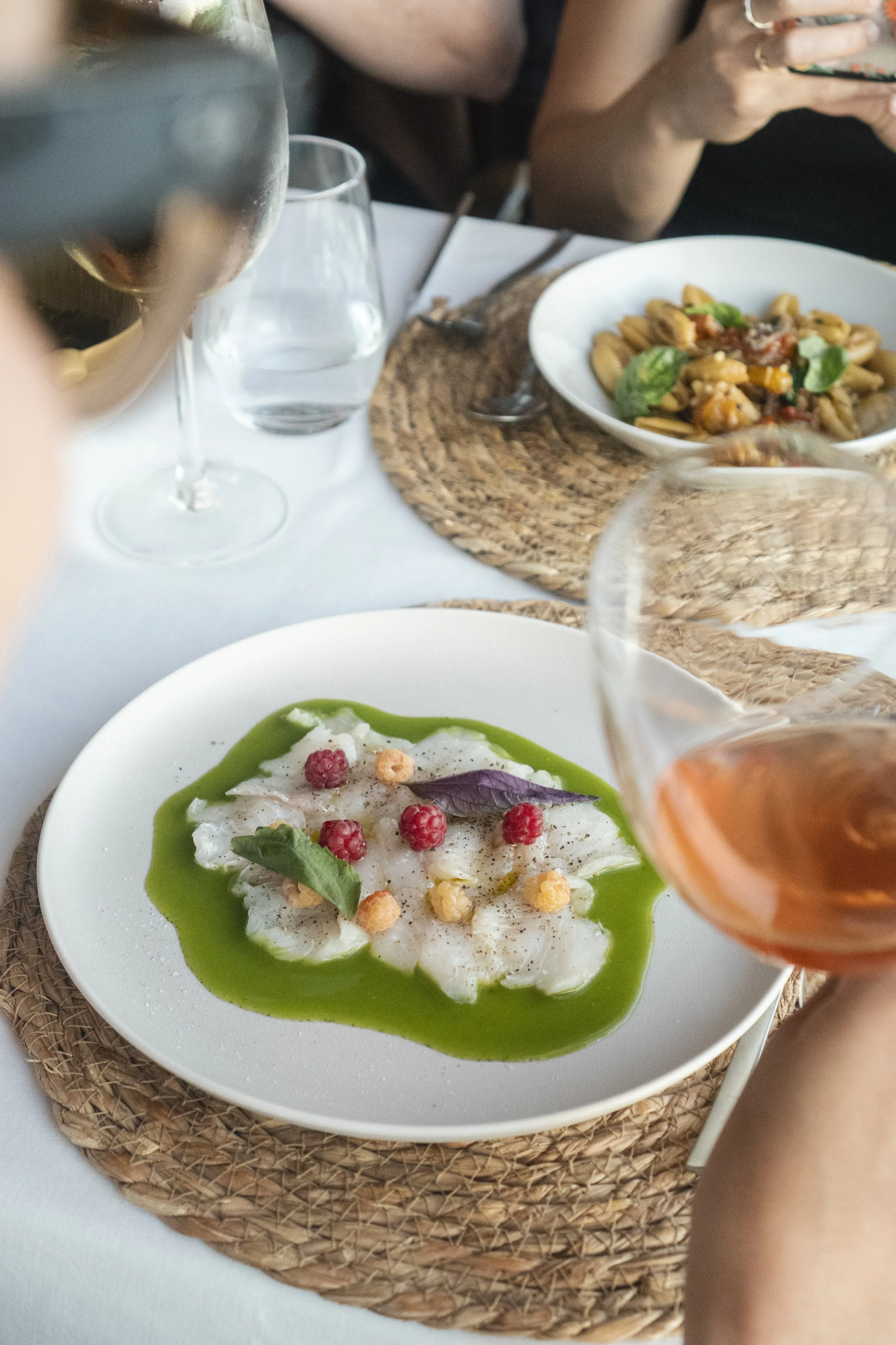Plato de ceviche adornado con frambuesas y hojas de albahaca, servido con salsa verde, en una mesa con vino rosado, agua y pasta en el fondo.