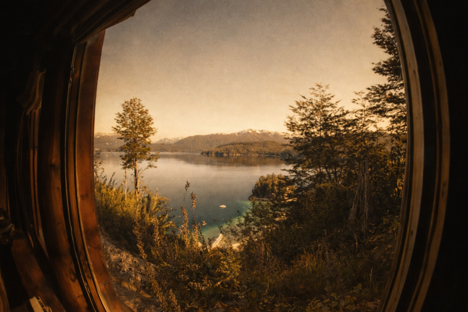 Vista de un lago con montañas al fondo, rodeado de árboles y vegetación, vista a través de una ventana redonda de madera.