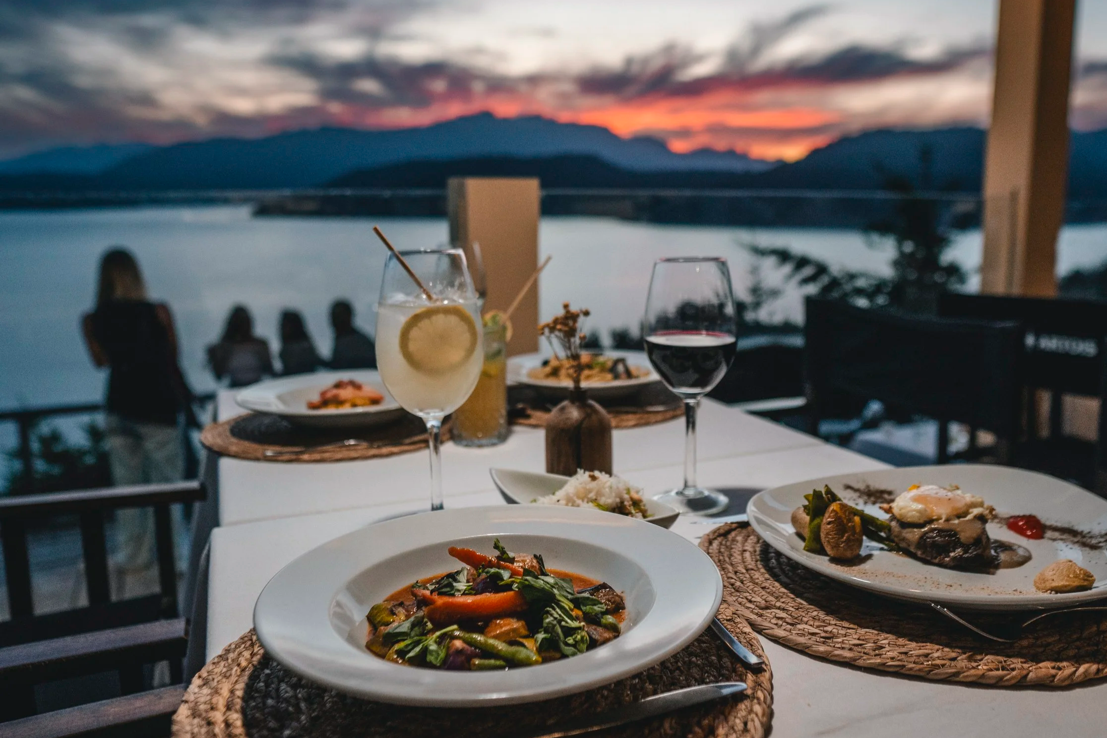 Cena en restaurante con vista al lago y montañas al atardecer, con platos de comida y bebidas en la mesa.