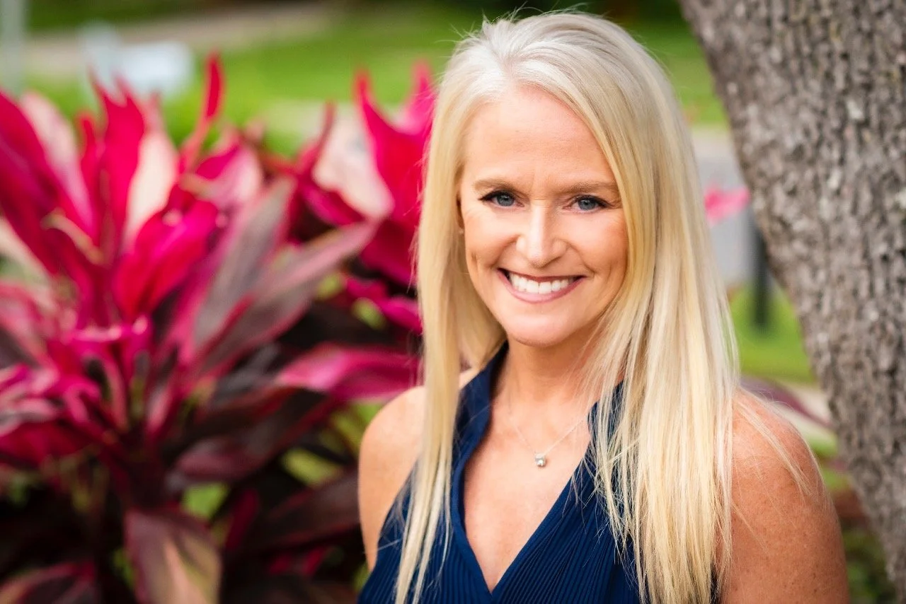 A smiling blonde woman in a blue sleeveless top standing outdoors with pink and green foliage in the background.