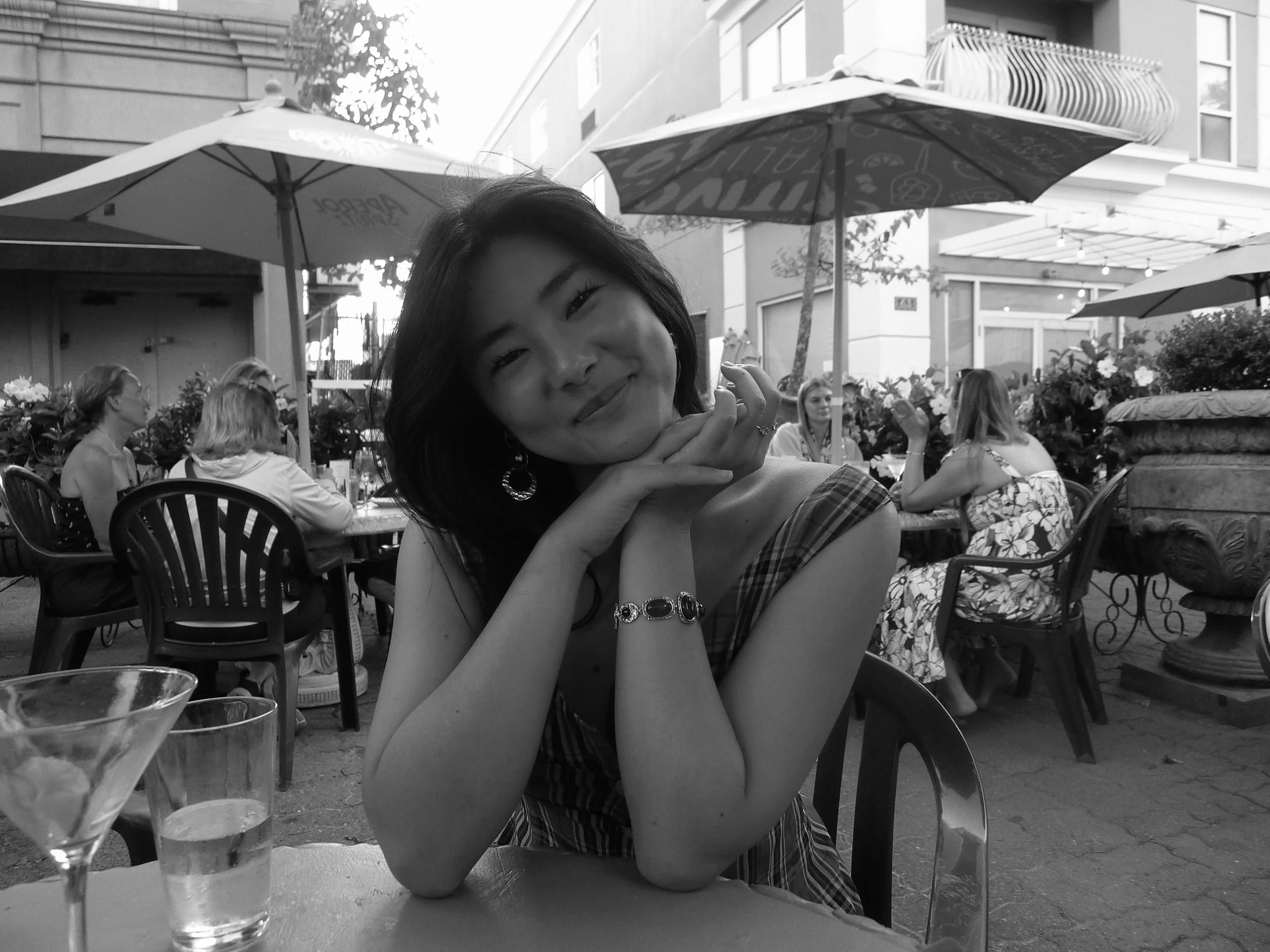 A young woman sitting at an outdoor cafe table, smiling with her head resting on her hands, surrounded by other diners and umbrellas, in a city setting.