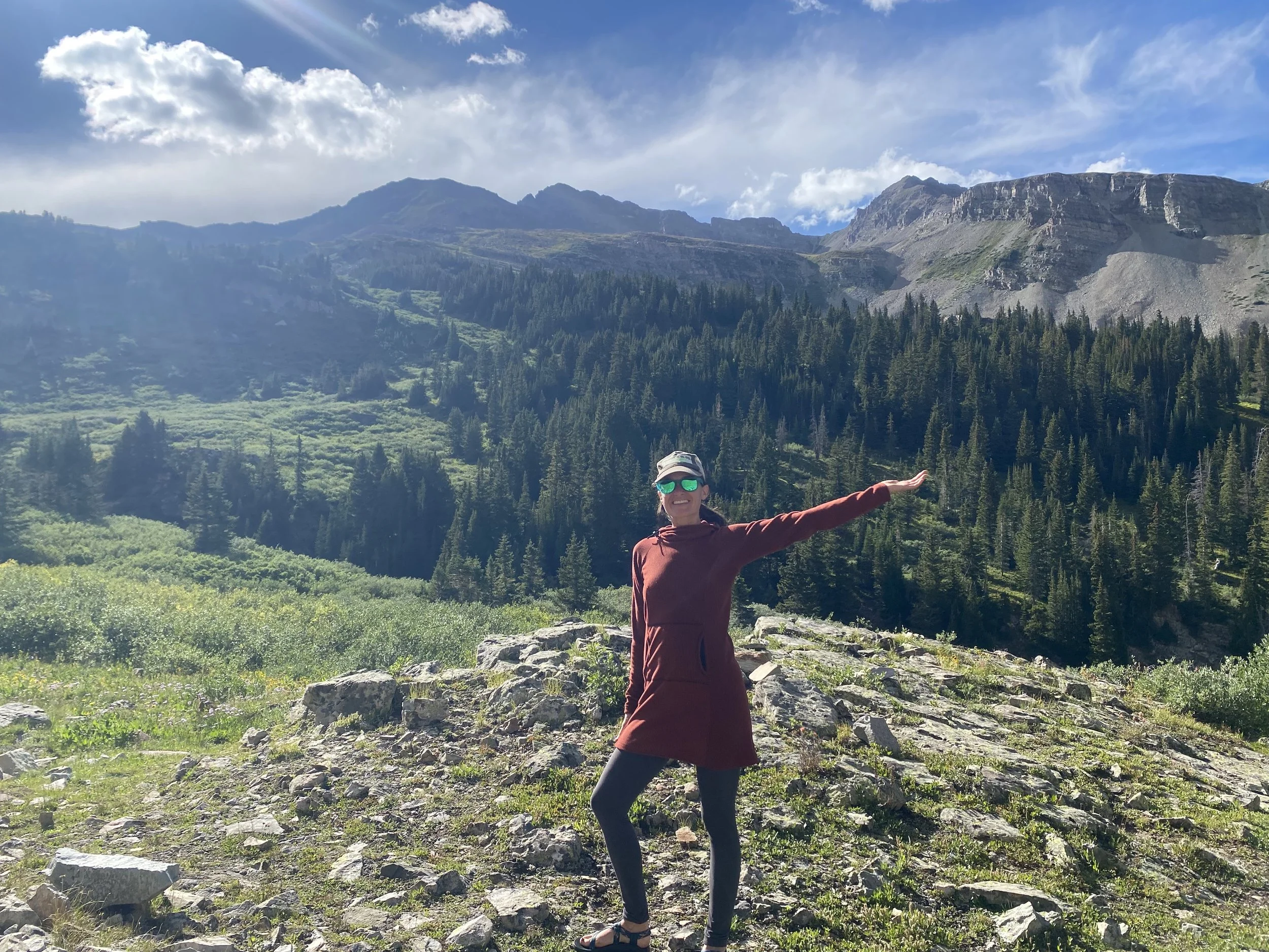 A woman wearing sunglasses, a cap, a maroon jacket, and black leggings stands on rocky terrain in a mountainous landscape with dense pine trees, green meadows, and distant mountain peaks, under a partly cloudy sky.