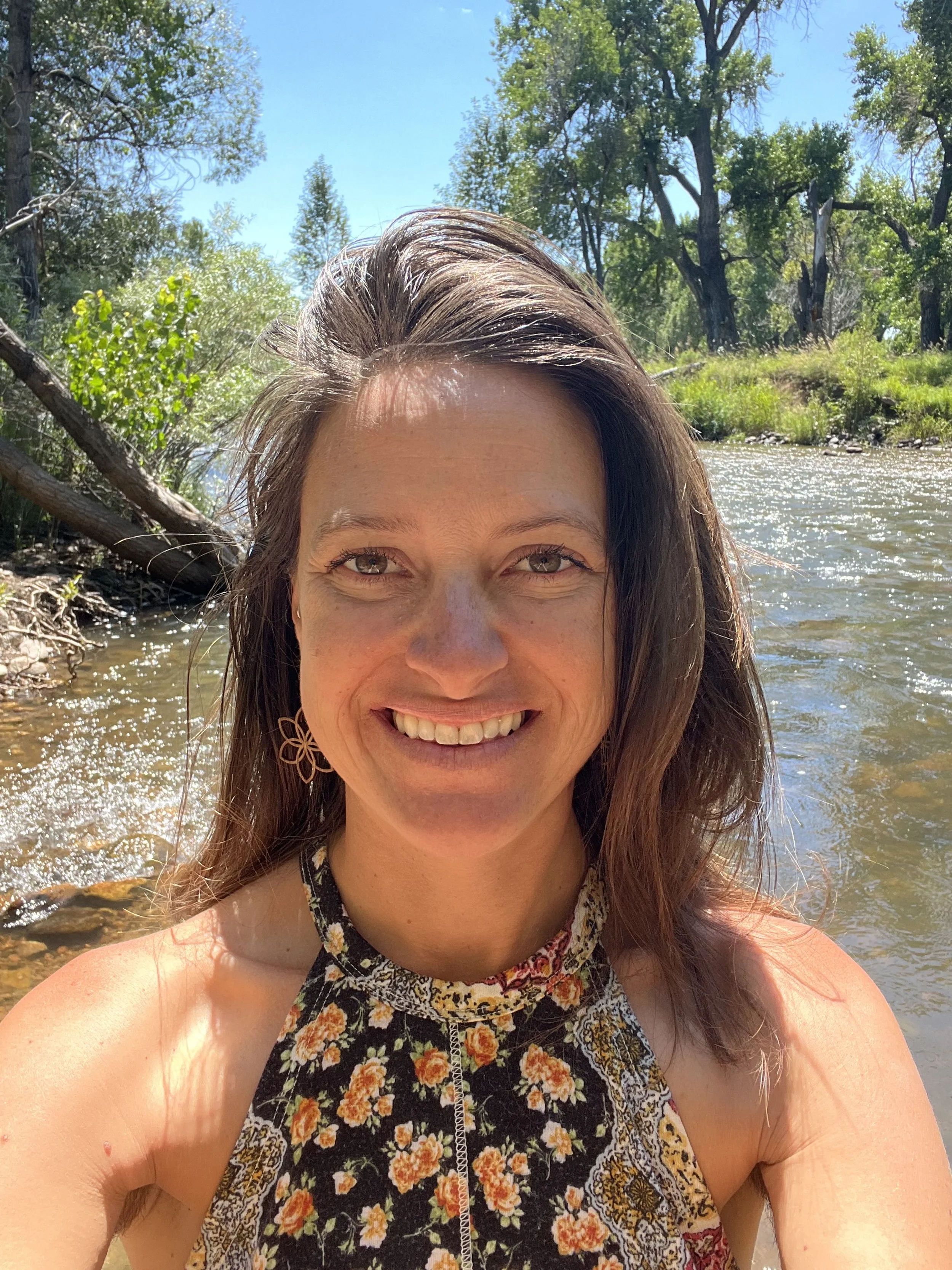 A woman smiling outdoors by a river with trees and a bright blue sky in the background.