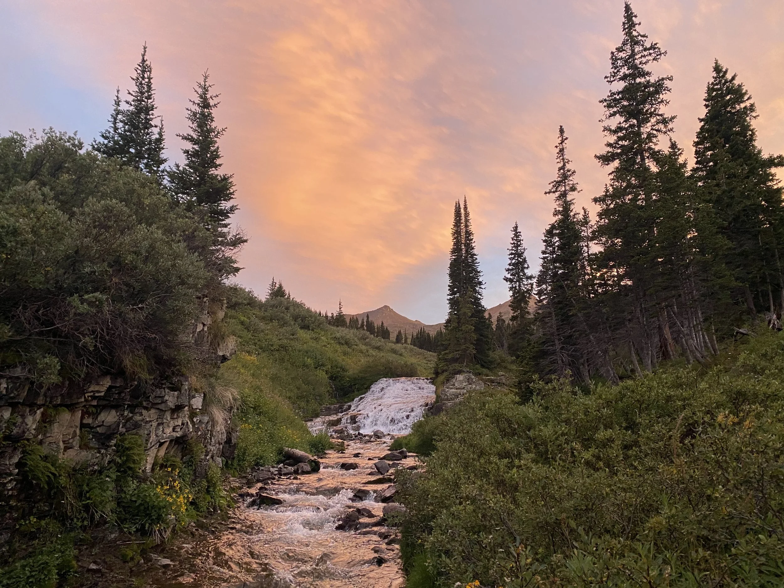 A mountain stream flowing through a lush forest under a colorful sky at sunset.