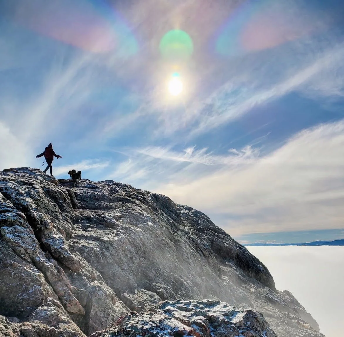 A person and a dog standing on rocky mountain edge with the sky and clouds behind them, the sun shining with lens flare effects.