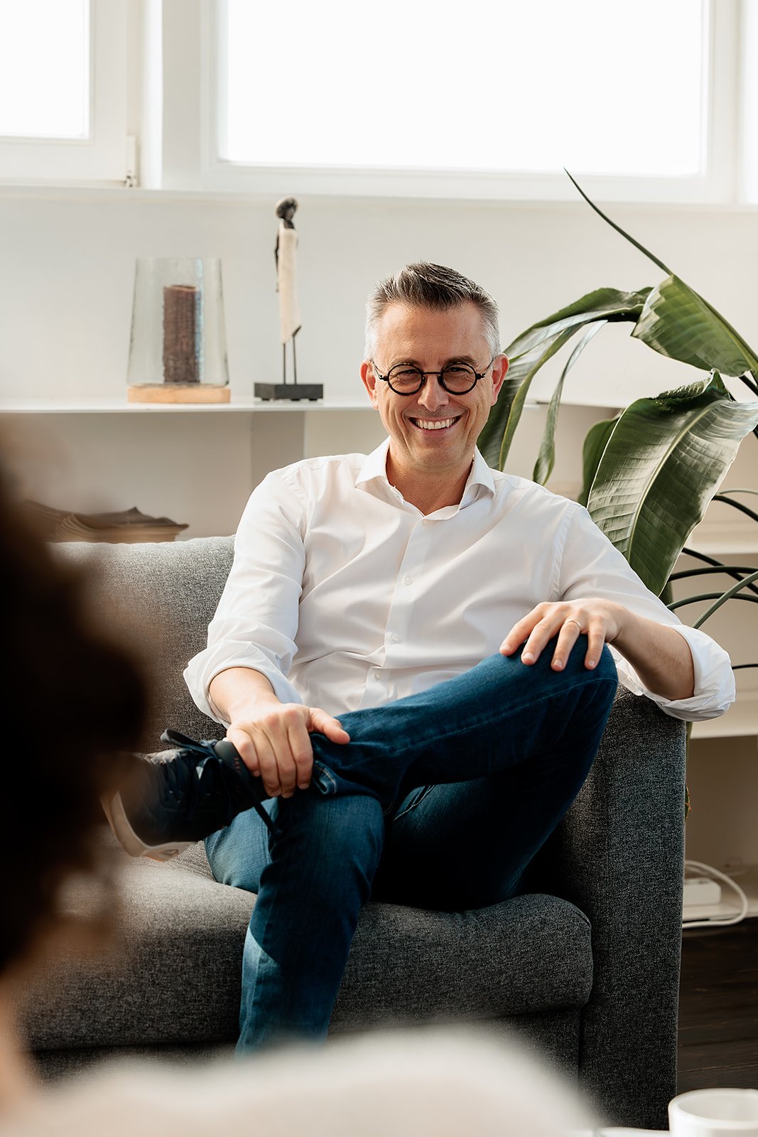 Man met bril zittend op een bank, lachend en ontspannen in een woonkamer met groene planten en decoratieve objecten op de achtergrond.