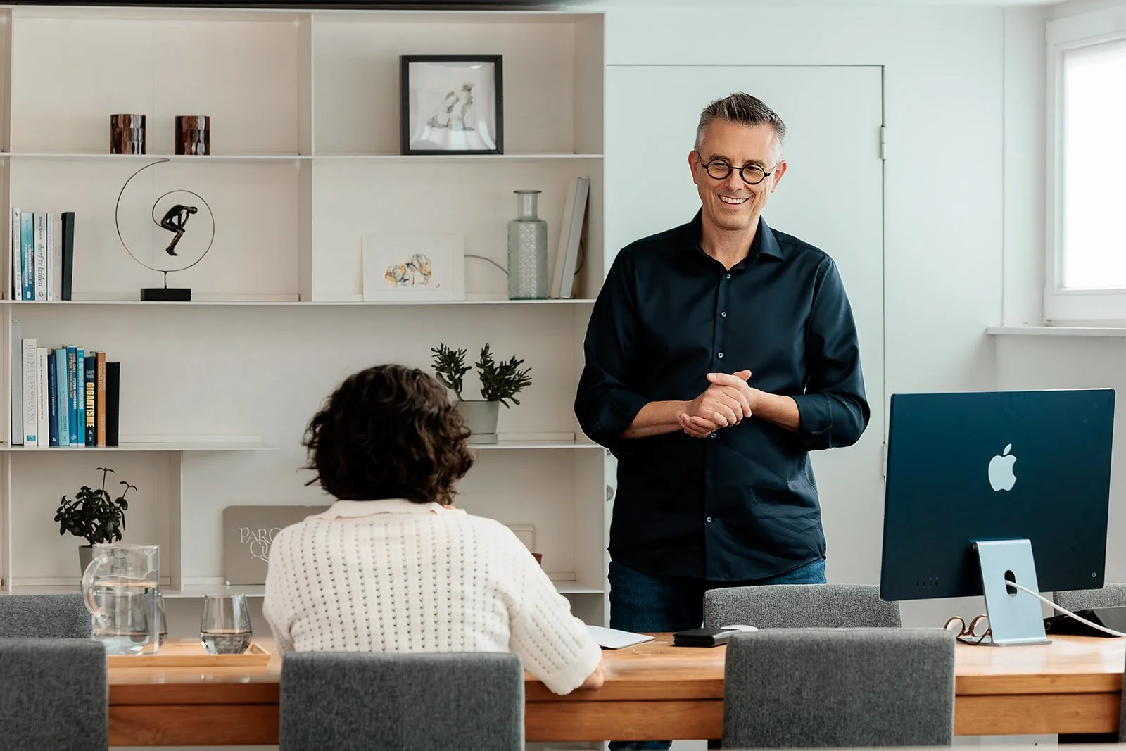 Mannelijke man staat voor een vrouw die aan een tafel zit in een vergaderruimte, met een computer en glazen water op de tafel, terwijl hij glimlacht.