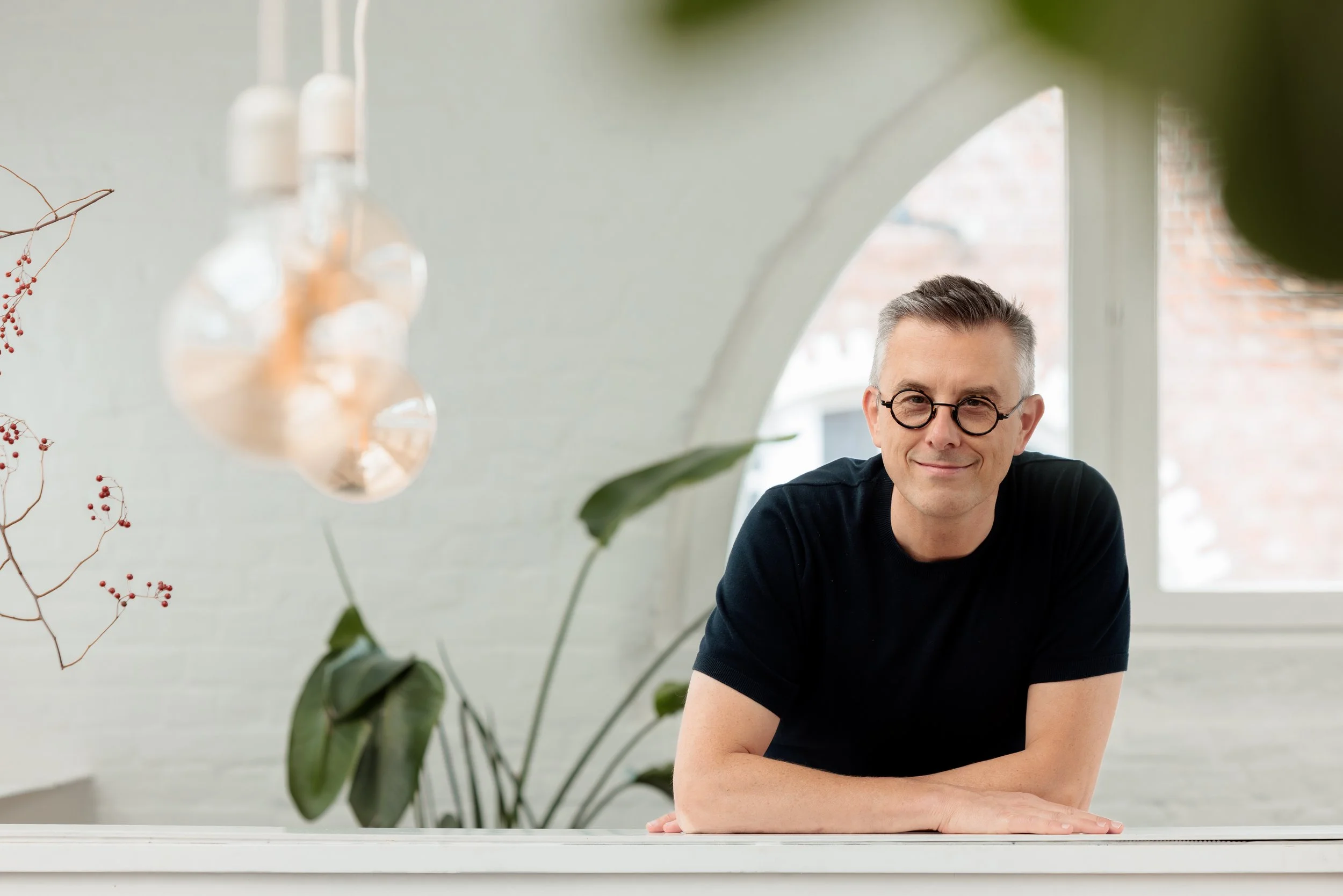 Man met bruin haar en ronde zwarte bril die glimlacht, zittend achter een witte tafel, met planten en een groot raam op de achtergrond.