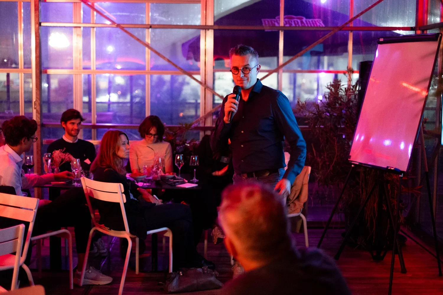 Een man met een microfoon spreekt voor een kleine groep mensen in een versierde zaal met ramen en roze/paars gekleurd licht.