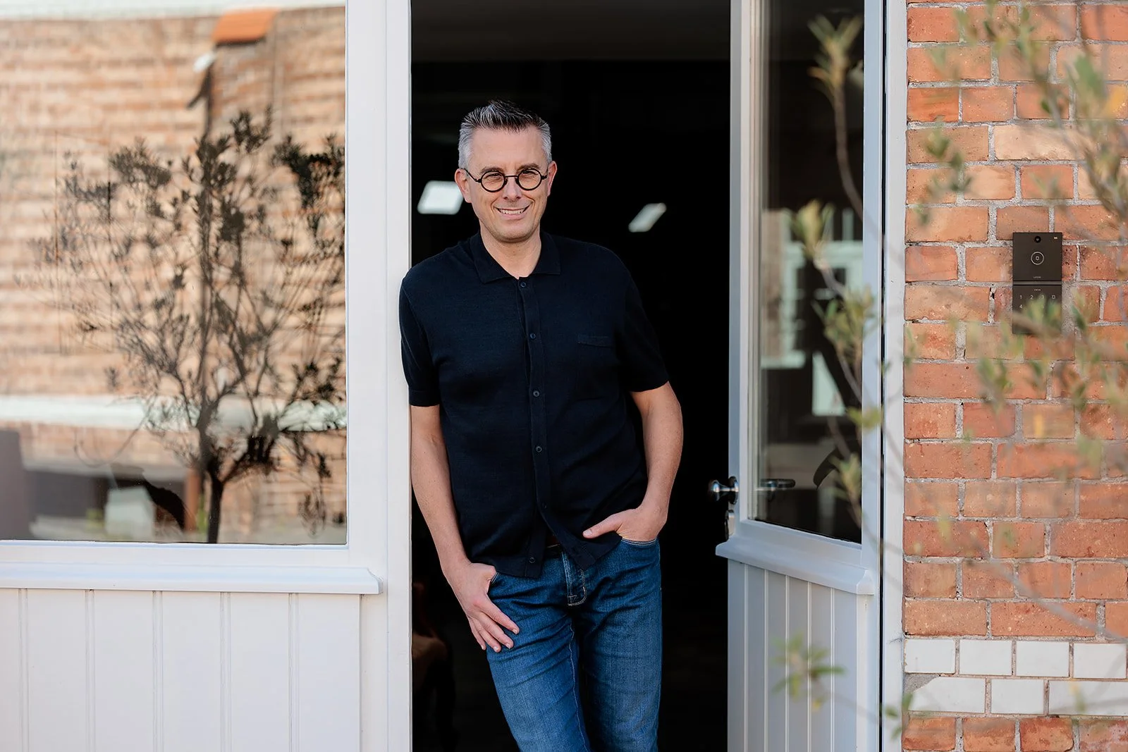 Man staat in de deuropening voor een huis, glimlacht naar de camera, draagt een zwart overhemd en blauwe jeans, achtergrond van huis en tuin.
