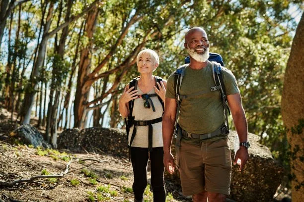 An older woman and man hiking on a forest trail, both carrying backpacks and smiling.