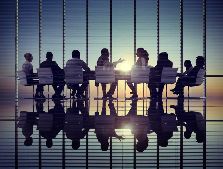 Business people in a meeting room having a discussion, sunlight outside through large windows, with reflections on the floor.