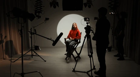 A woman with long hair sitting in a studio with professional filming equipment, including cameras and lighting, as she is being recorded or photographed.