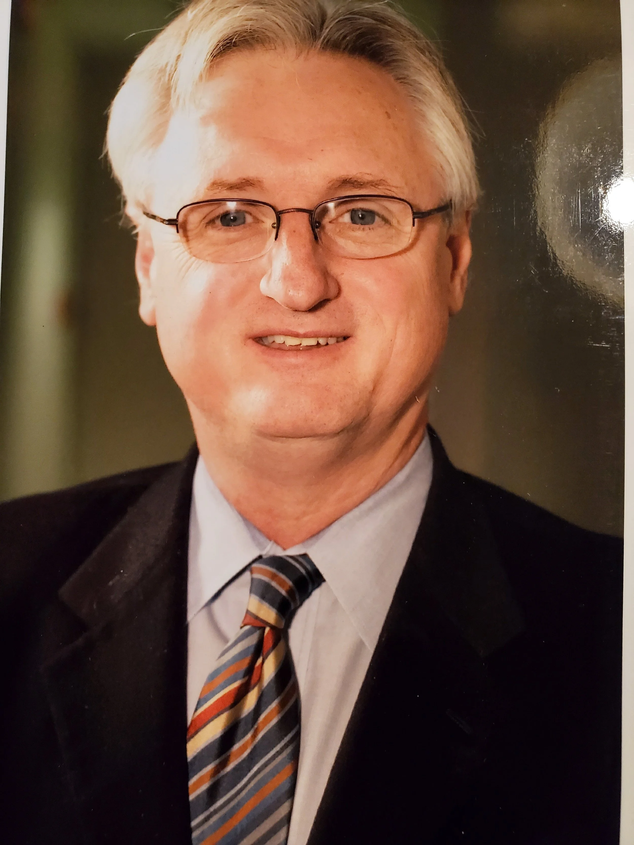A middle-aged man with glasses, blond hair, wearing a black suit, a light-colored dress shirt, and a striped tie, smiling at the camera.