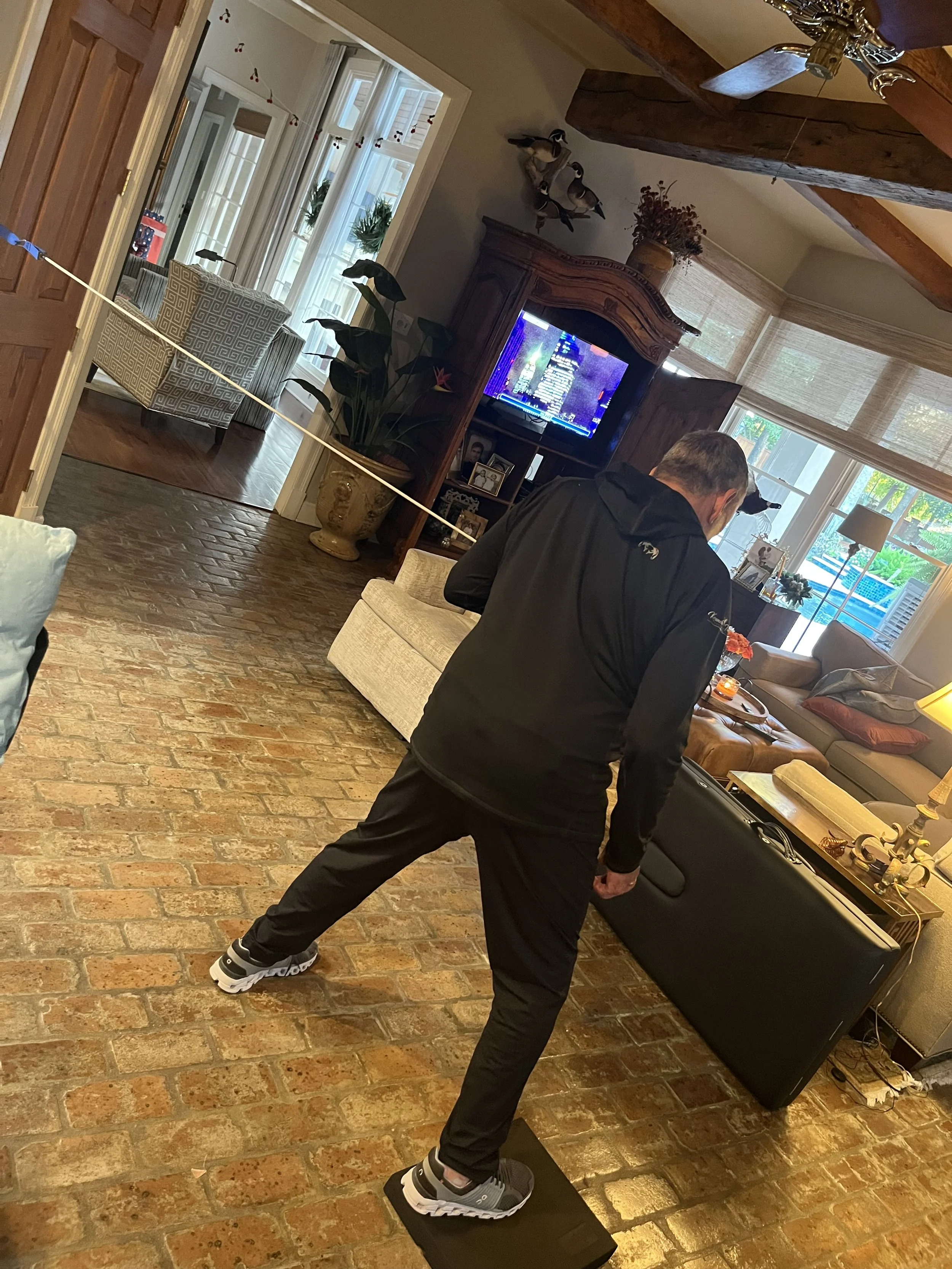 A man in a black jacket and pants is standing on a black step platform doing excercise in a living room, facing away from the camera. The room has brick flooring, sofas, a TV, and large windows with sunlight streaming in.